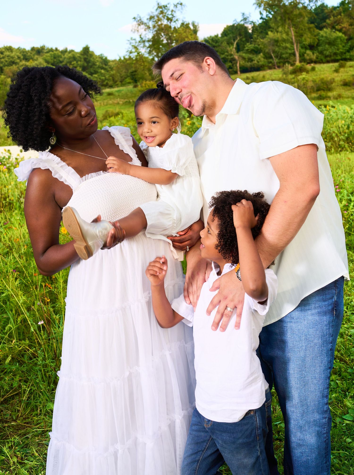Happy interracial family in white outfits posing outdoors in a lush green field during a portrait session.