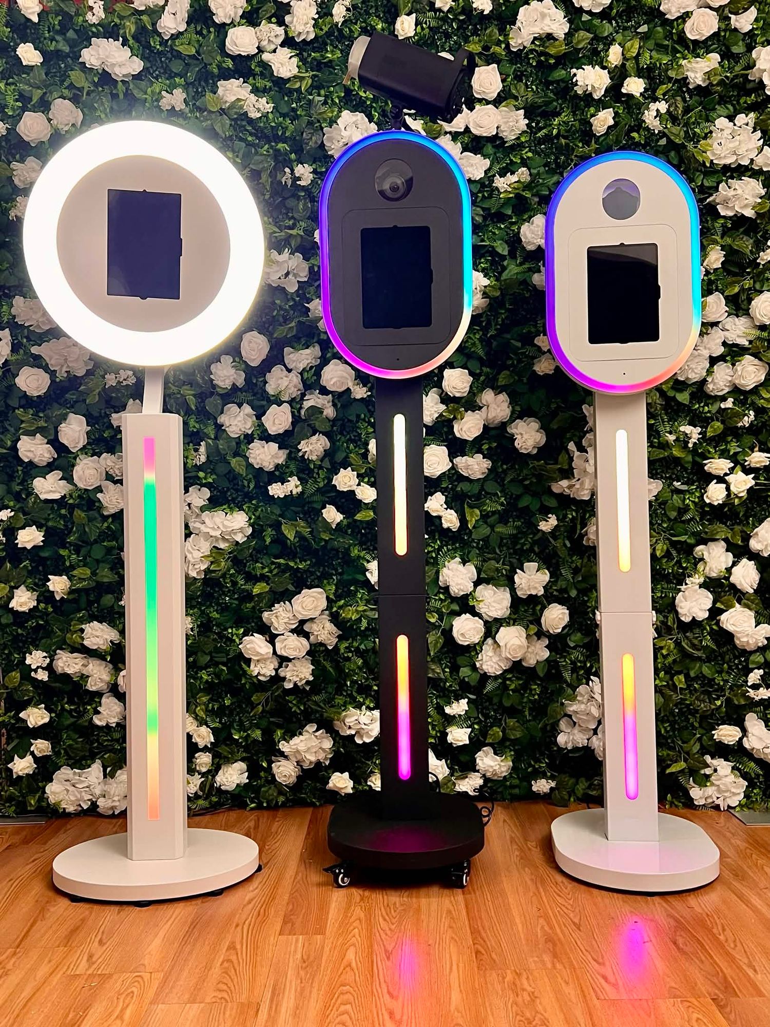 Three modern photo booth setups with RGB lighting against a white floral wall, including ring light and 360 platforms.