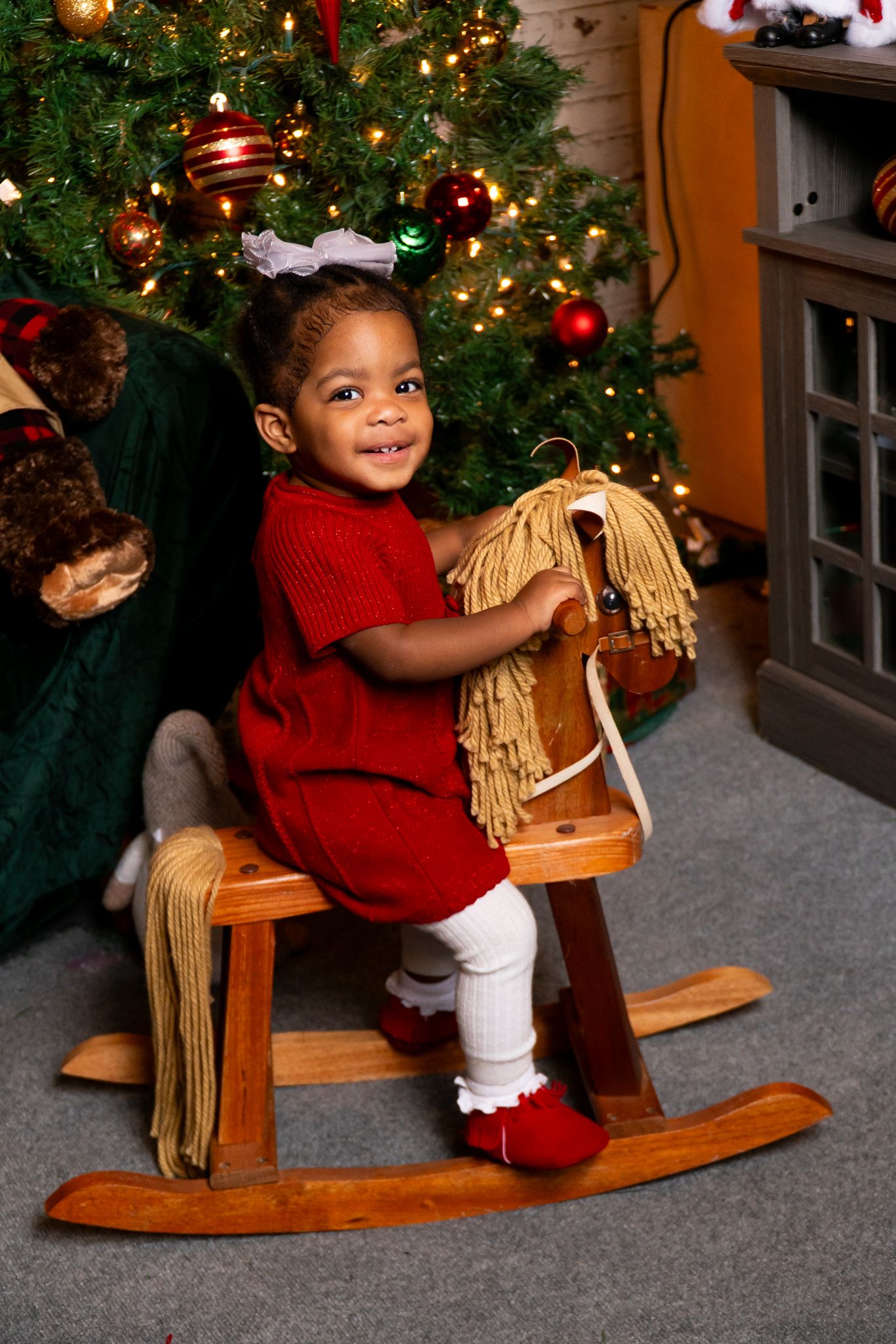 Adorable toddler in red dress smiling while riding a wooden rocking horse by a Christmas tree.