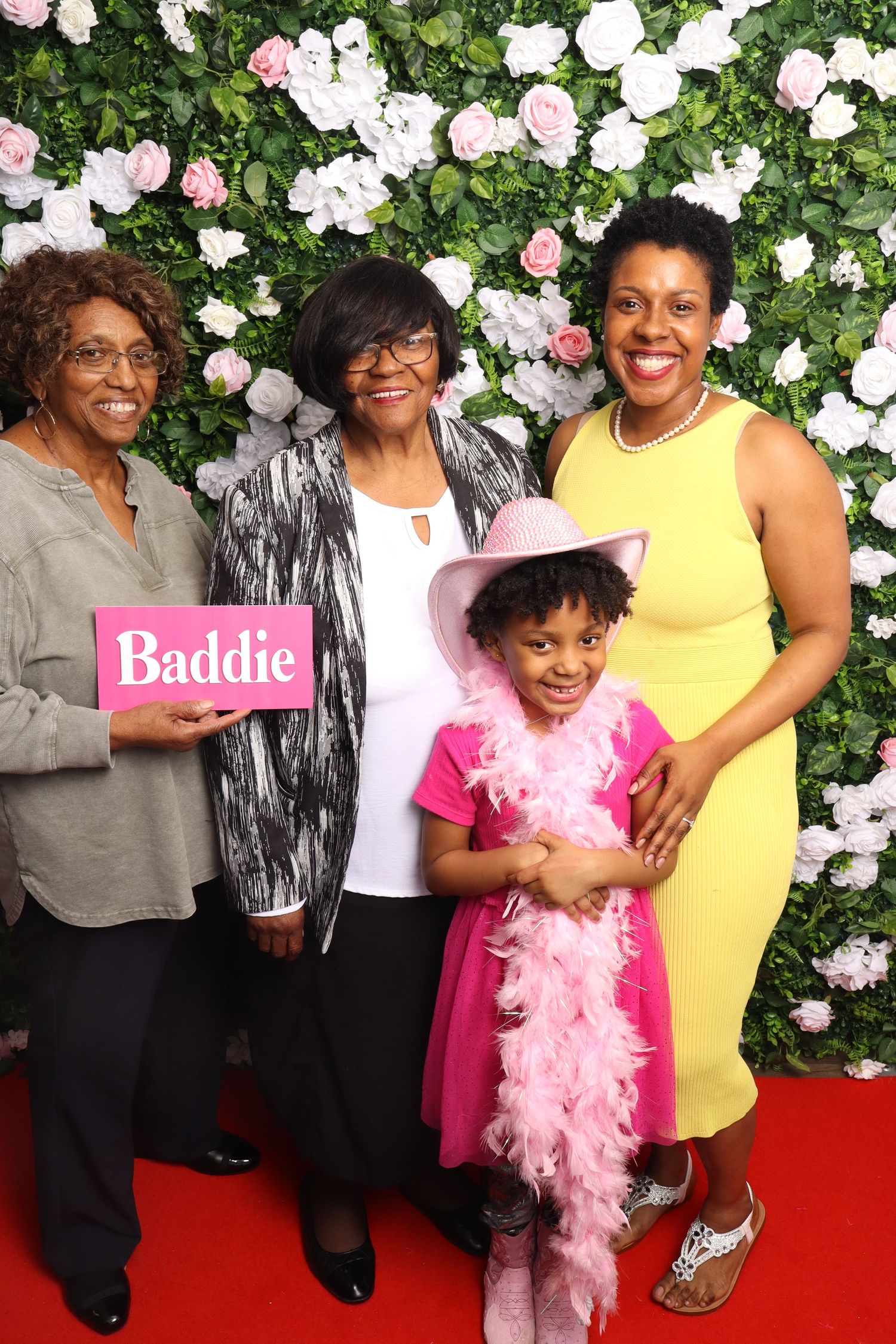 Three women and a young girl in pink pose with a Baddie sign in front of a floral wall backdrop.