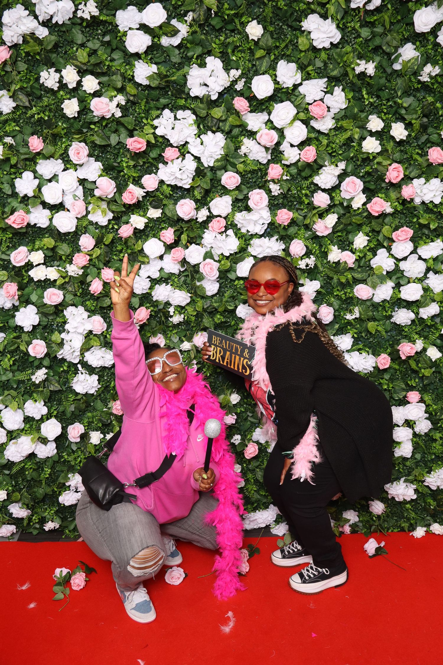 Two people in colorful pink outfits pose playfully in front of a flower wall backdrop on a red carpet.