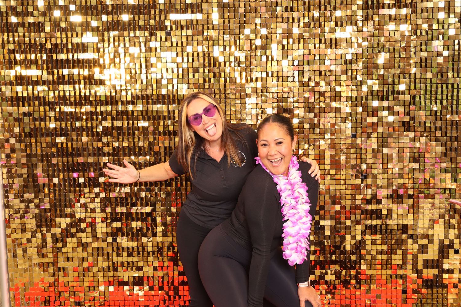 Two women posing and smiling in front of a gold sequin backdrop, one wearing sunglasses and a lei.