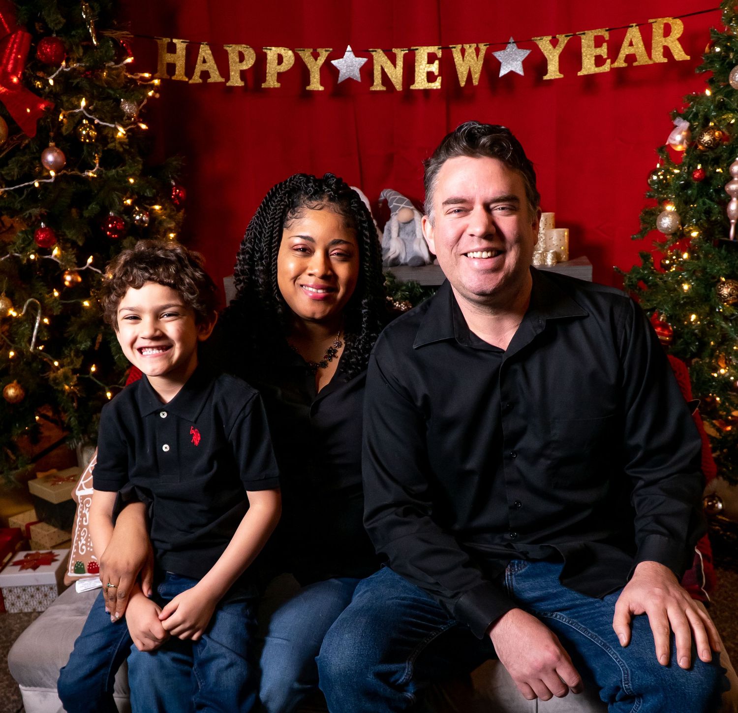Smiling family of three poses together under a Happy New Year banner with a decorated Christmas tree.