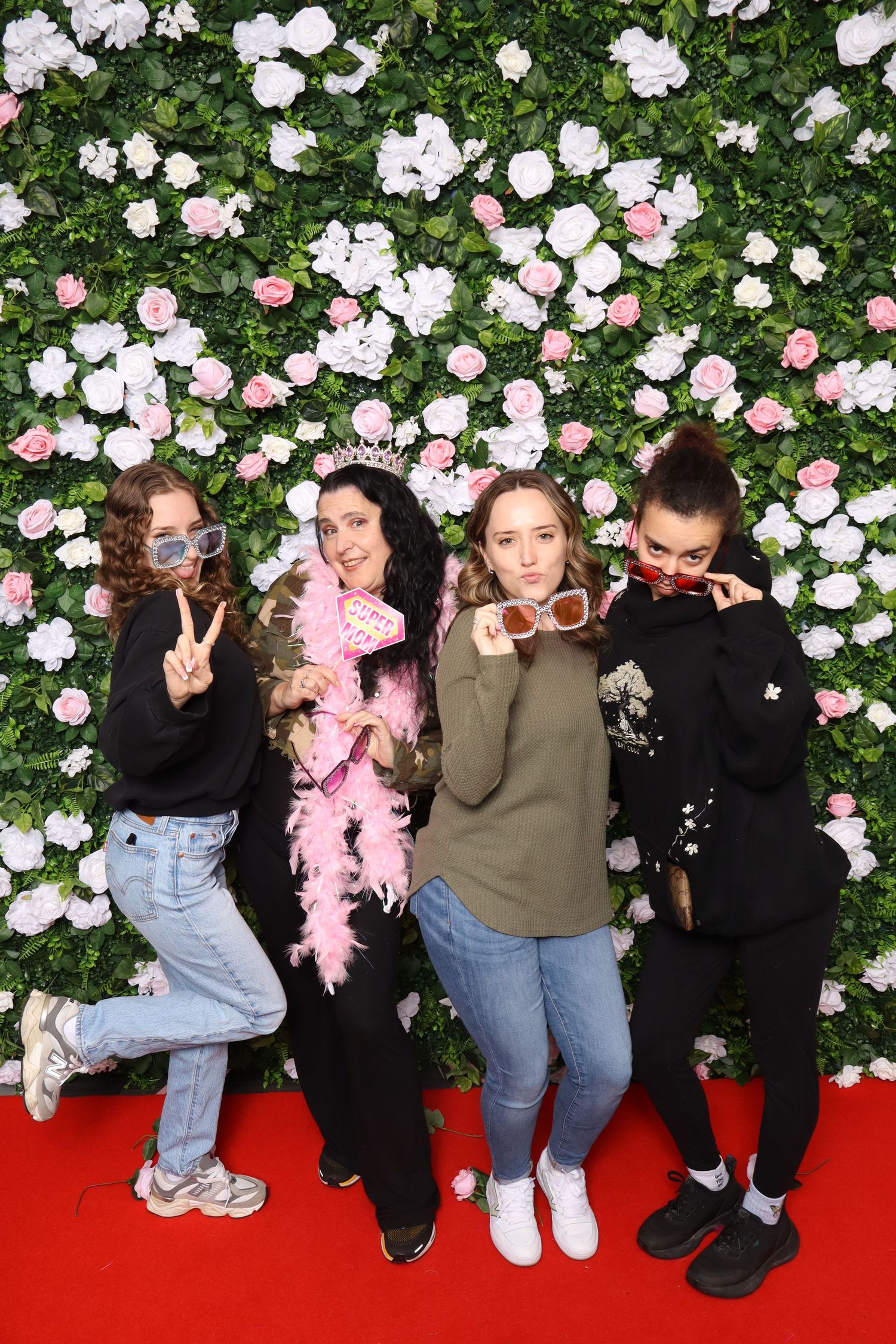 Four young women posing in front of a floral wall backdrop on a red carpet, wearing fun accessories.