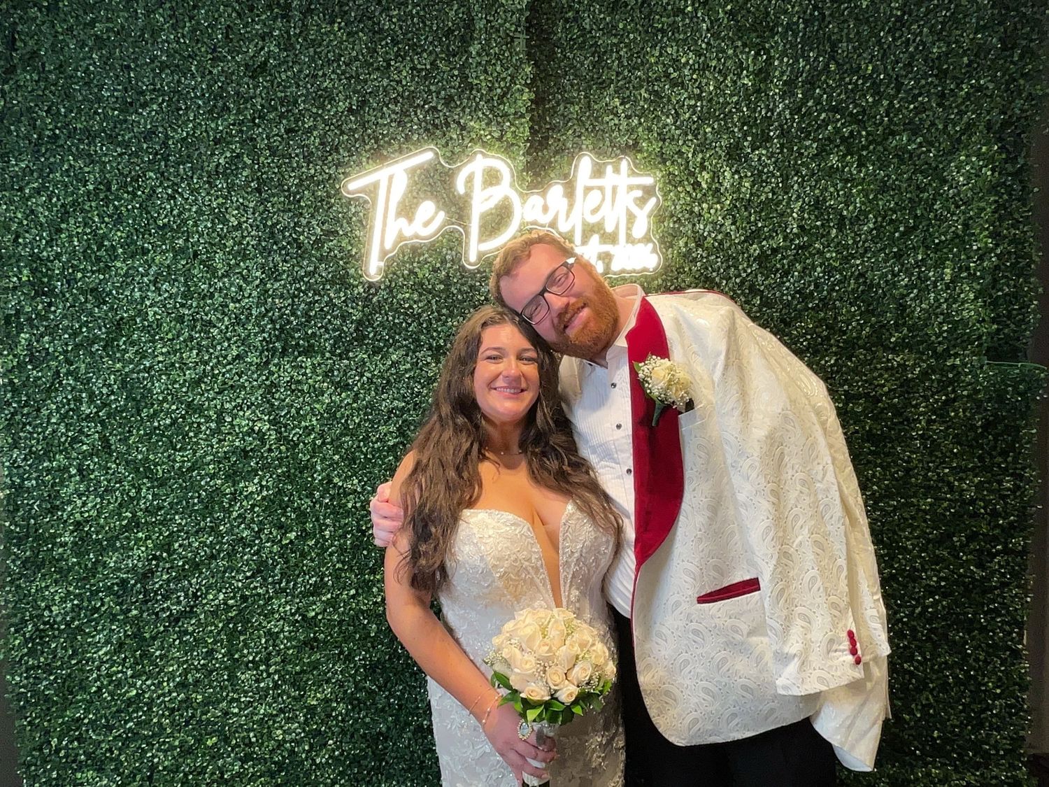Newlywed couple posing in front of a green hedge wall with The Bartlett neon sign at their wedding reception.