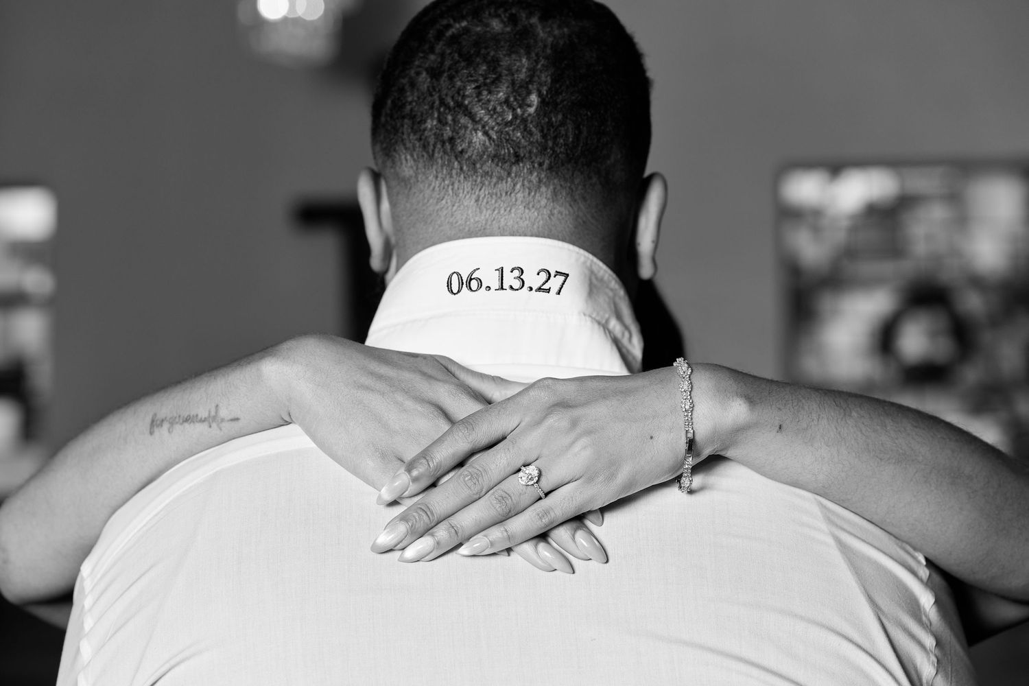Black and white photo of a couple embracing, showing a tattoo reading 06.13.27 on the mans neck.