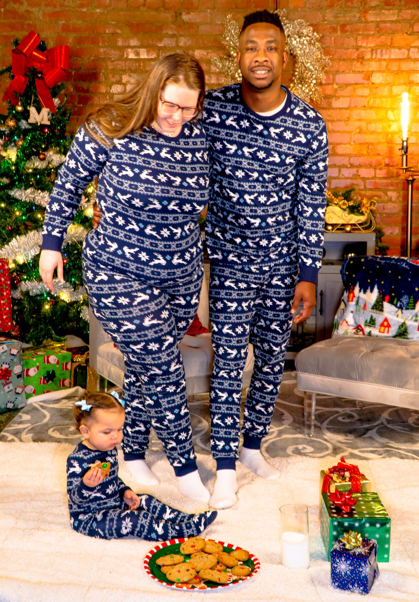 Family in matching navy blue Fair Isle Christmas pajamas stands near tree with plate of cookies.