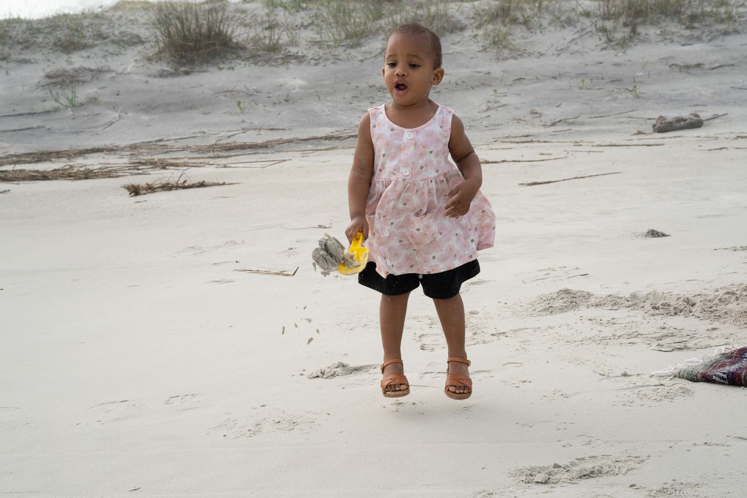 Small figure wearing a white sleeveless top runs joyfully on sandy beach while holding yellow toy.