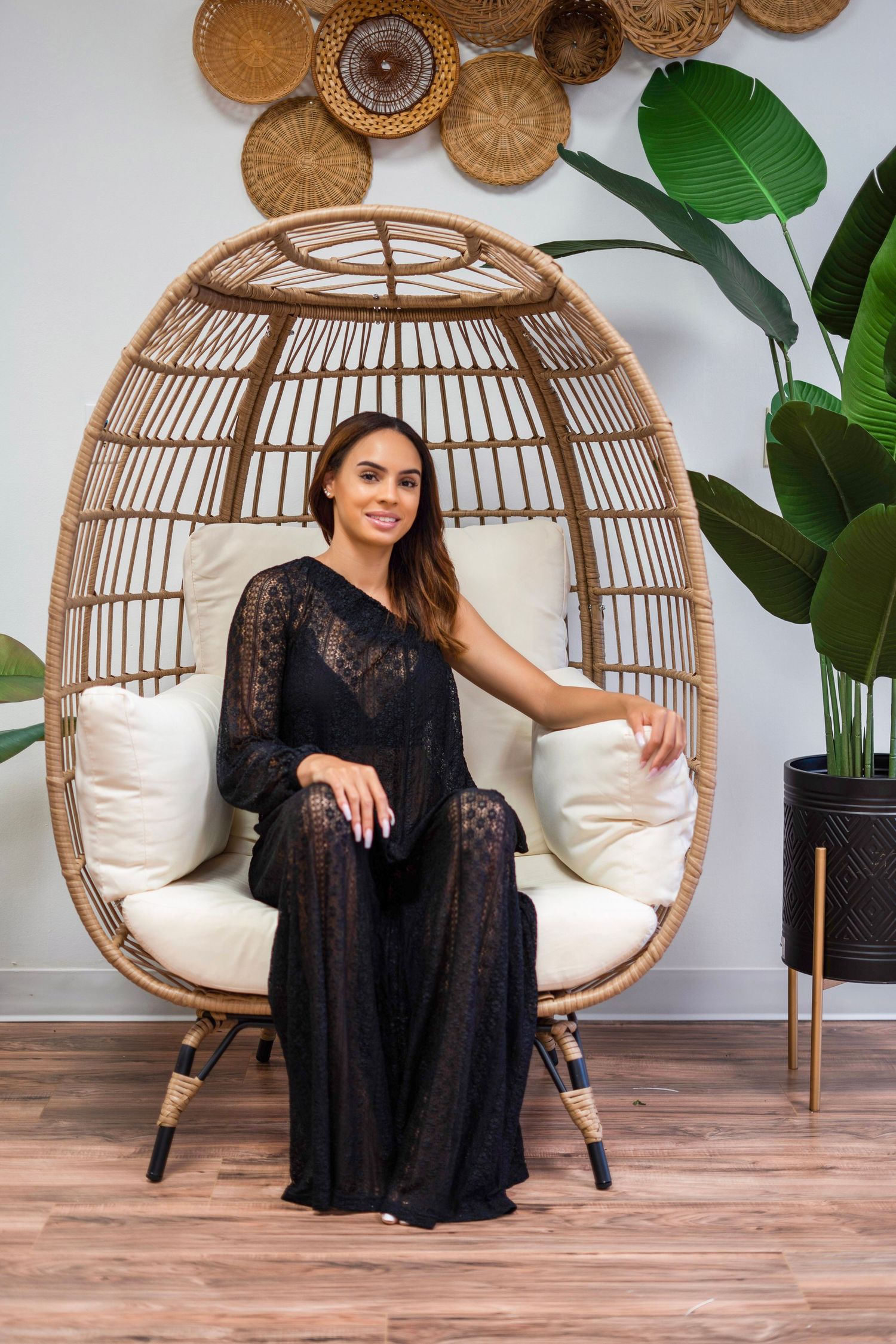 A person in a black maxi dress poses in a hanging rattan egg chair with decorative wall decor and plants.