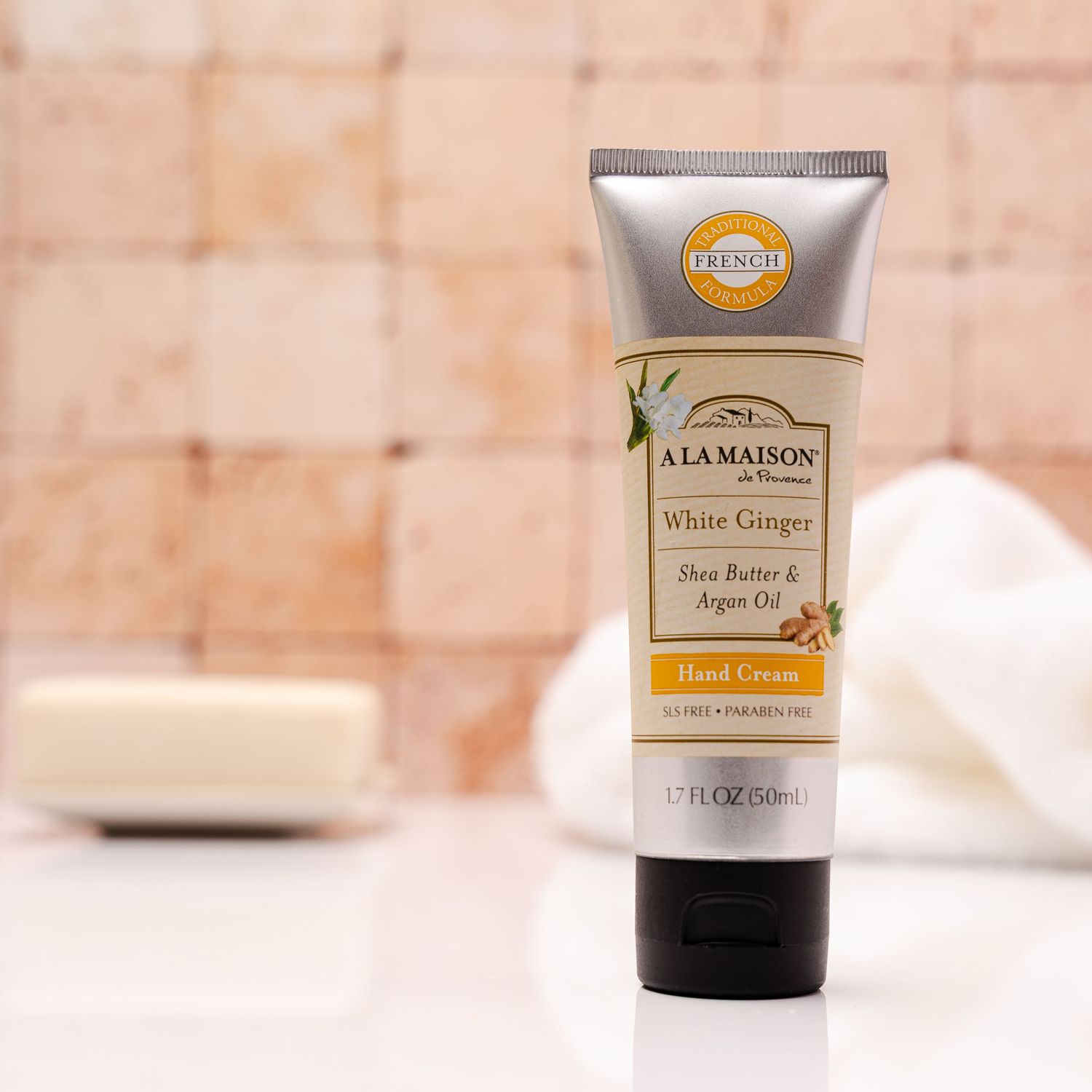 A tube of A La Maison White Ginger hand cream sits against a blurred beige background with a white bar in the distance.