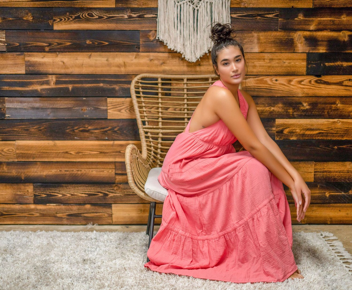 Person in coral maxi dress poses on rattan chair against wooden wall with macrame decor.