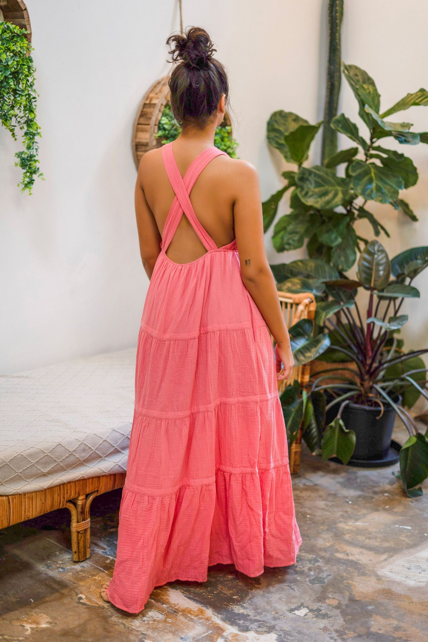 Back view of coral maxi dress showing criss-cross straps and tiered design among indoor plants.