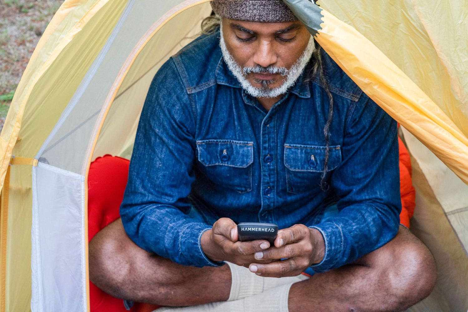 Person in denim jacket looking down at mobile phone while sitting in front of yellow tent.