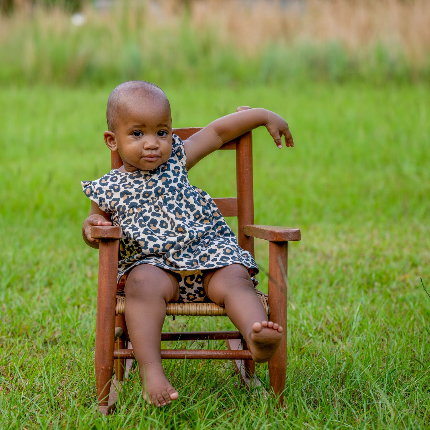 A small child wearing a leopard print dress sits in a wooden rocking chair on green grass.