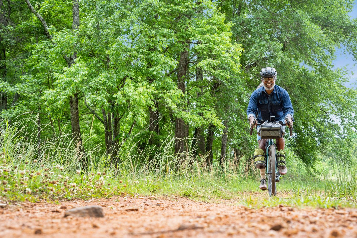 Mountain biker rides through a wooded trail with green foliage and reddish dirt path in a sequence of action shots.