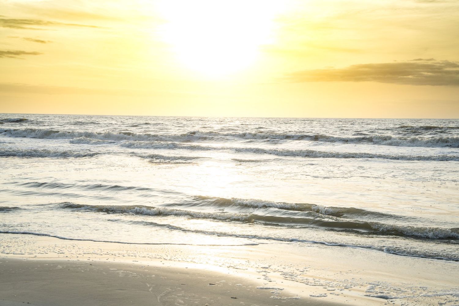 Golden sunset over a serene beach with gentle waves rolling onto shore and soft light reflecting on the ocean surface.