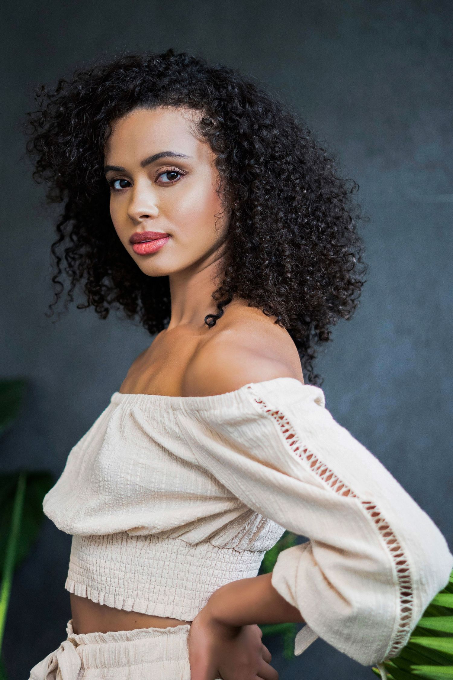 A model in an off-shoulder cream top poses against a dark background with tropical leaves.
