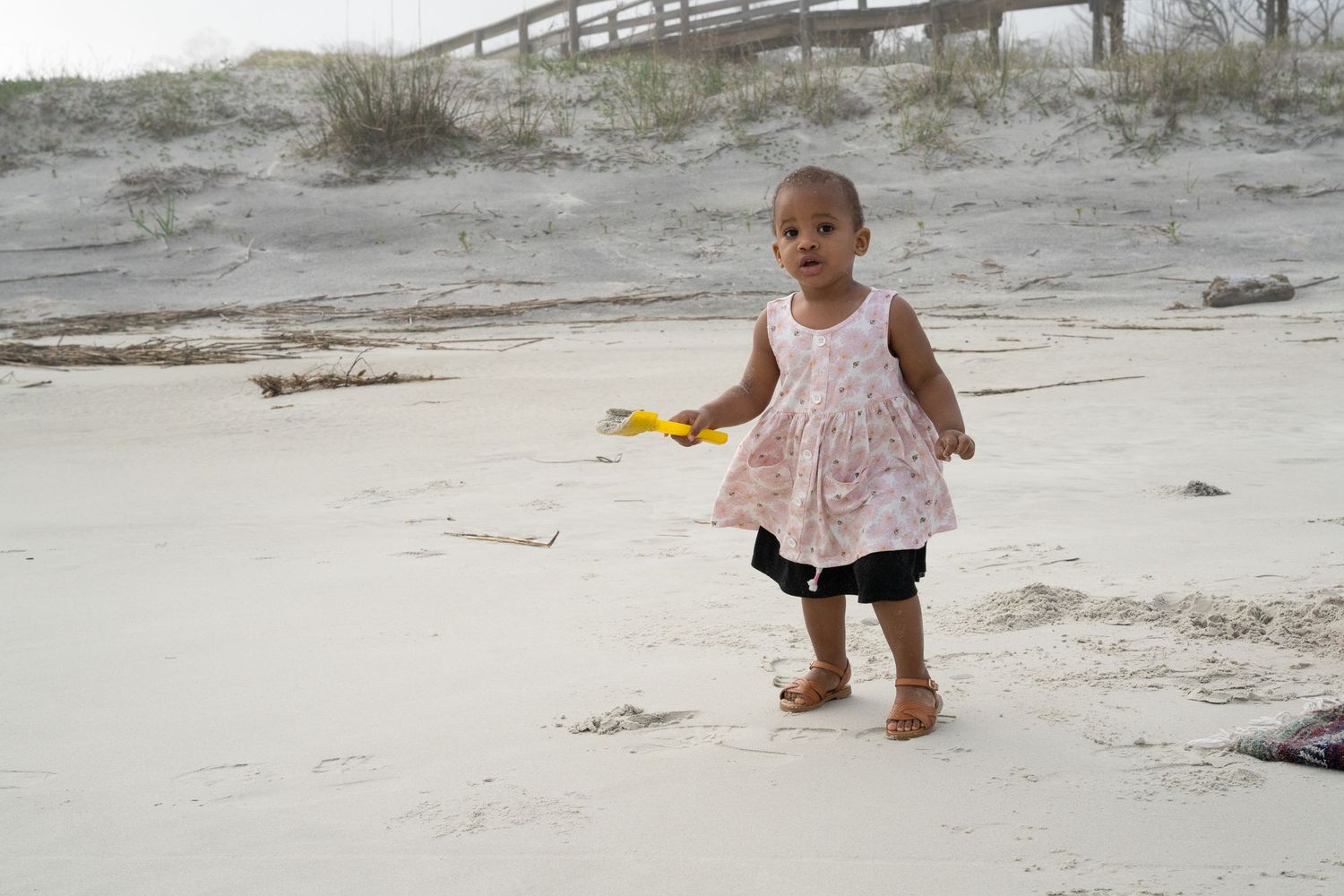 Small toddler wearing a pink dress plays with a yellow toy while standing barefoot on a sandy beach near a wooden boardwalk.