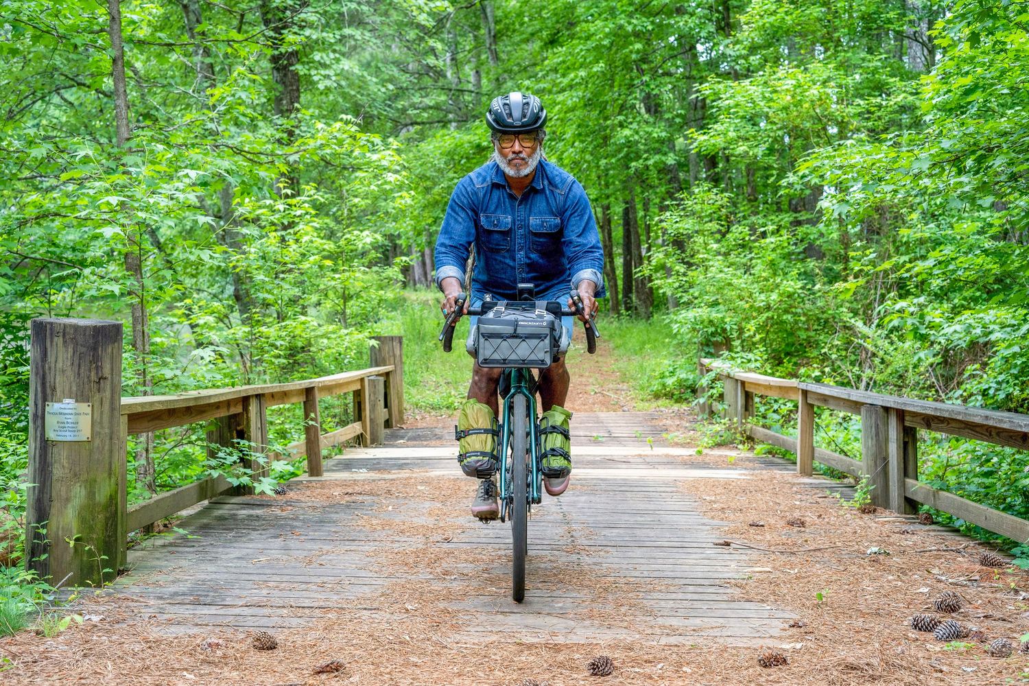 A cyclist rides on a wooden boardwalk through lush green forest with bike panniers on a scenic trail.