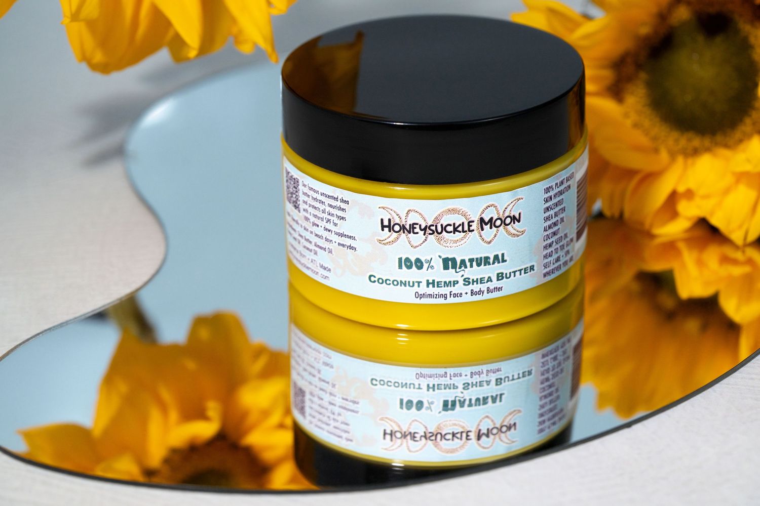 A black-lidded jar of hair treatment product displayed on a mirror surface with yellow sunflowers in the background.