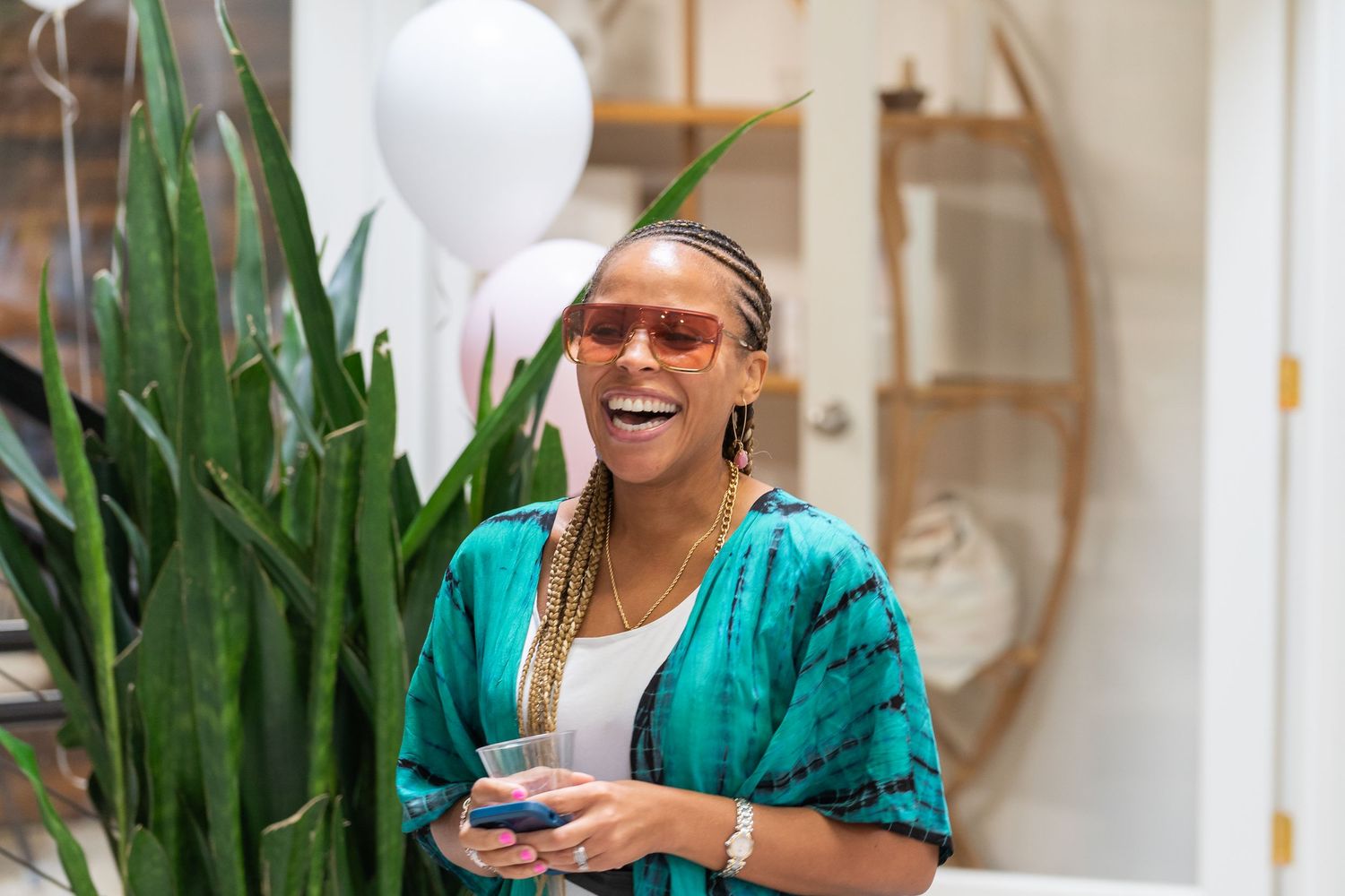 Someone wearing a turquoise jacket and sunglasses laughs joyfully while holding a drink near an indoor plant.