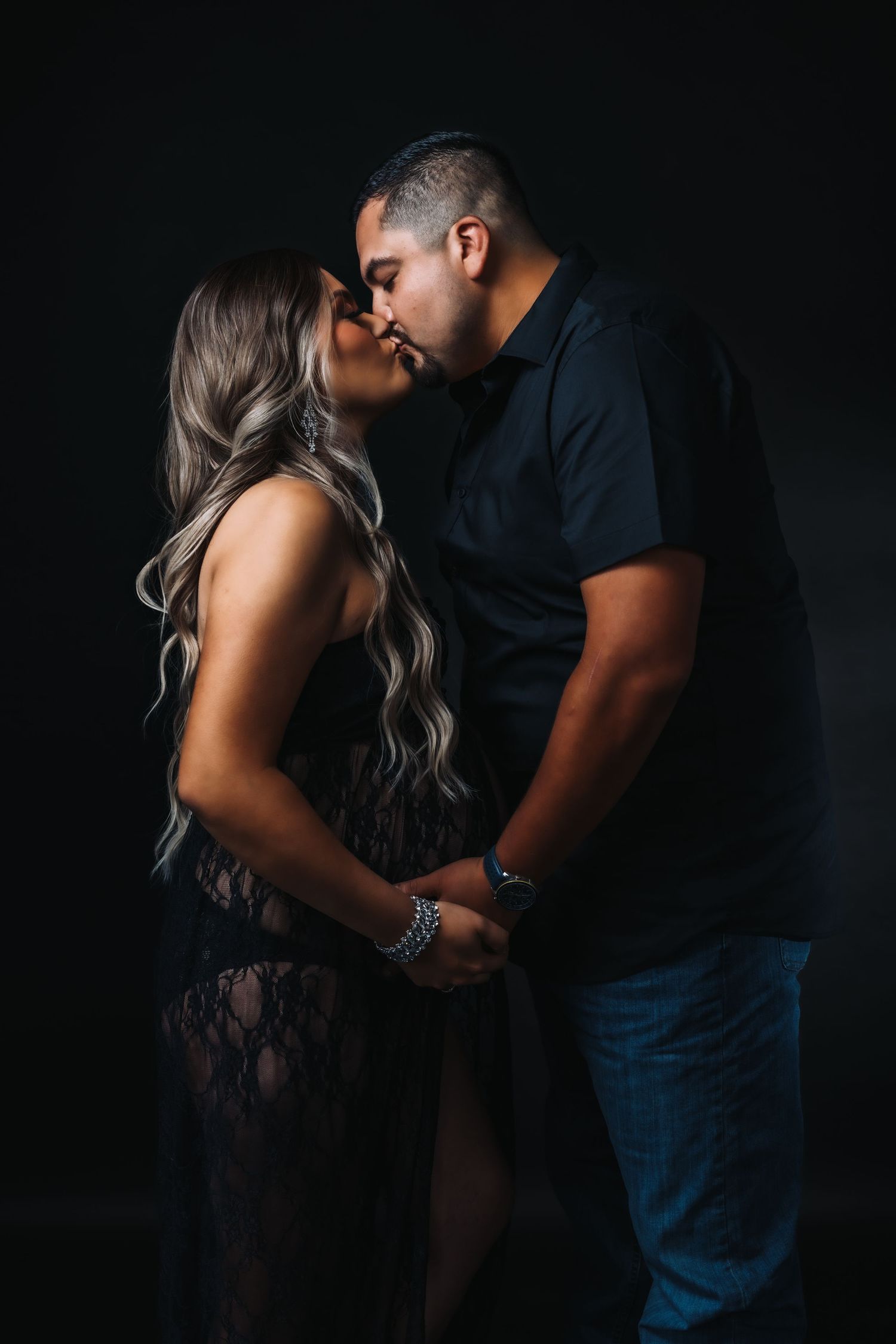 Intimate maternity couple photoshoot in dark studio setting with dramatic lighting.