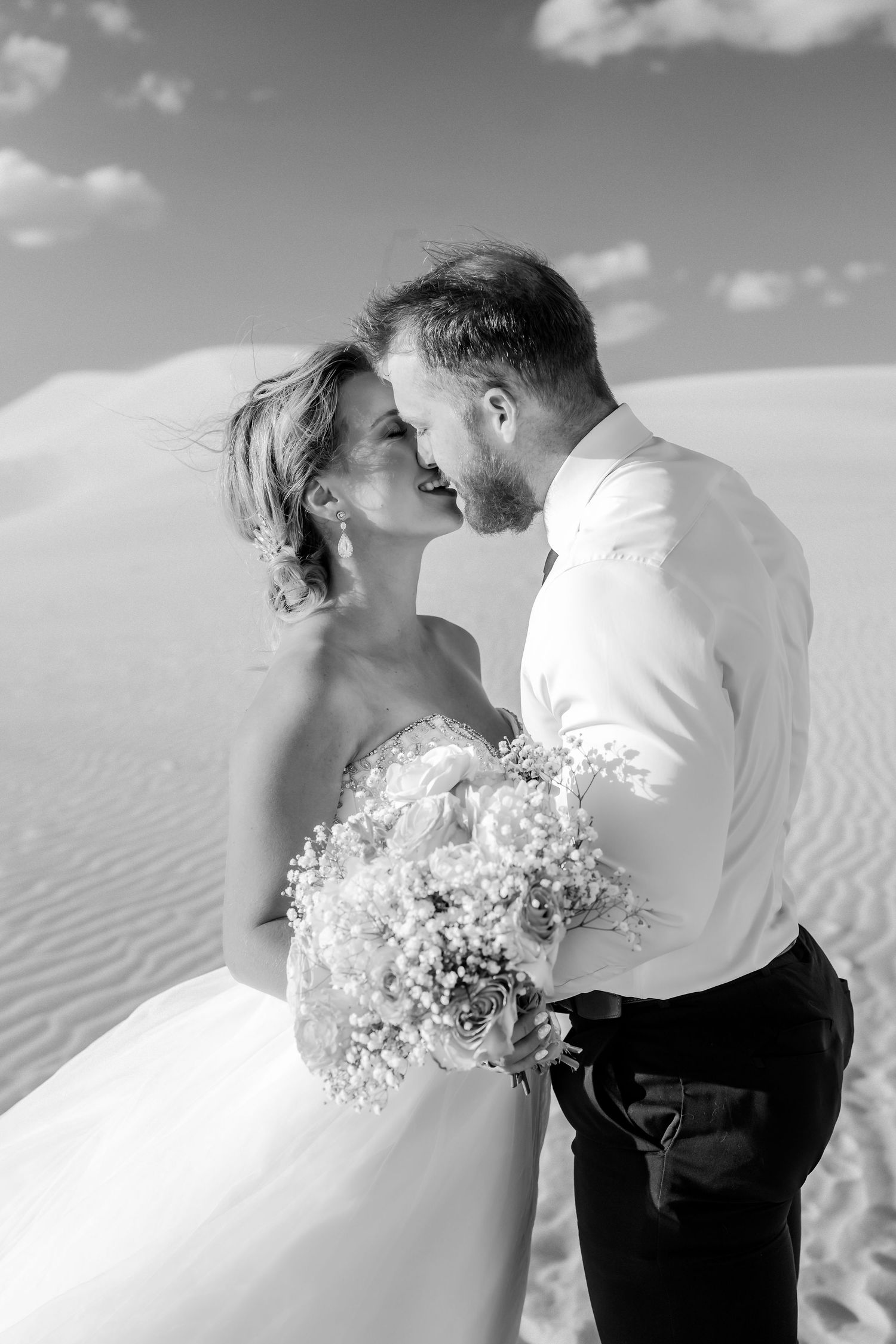 Romantic couple embraces in white attire against sandy dunes under cloudy sky.