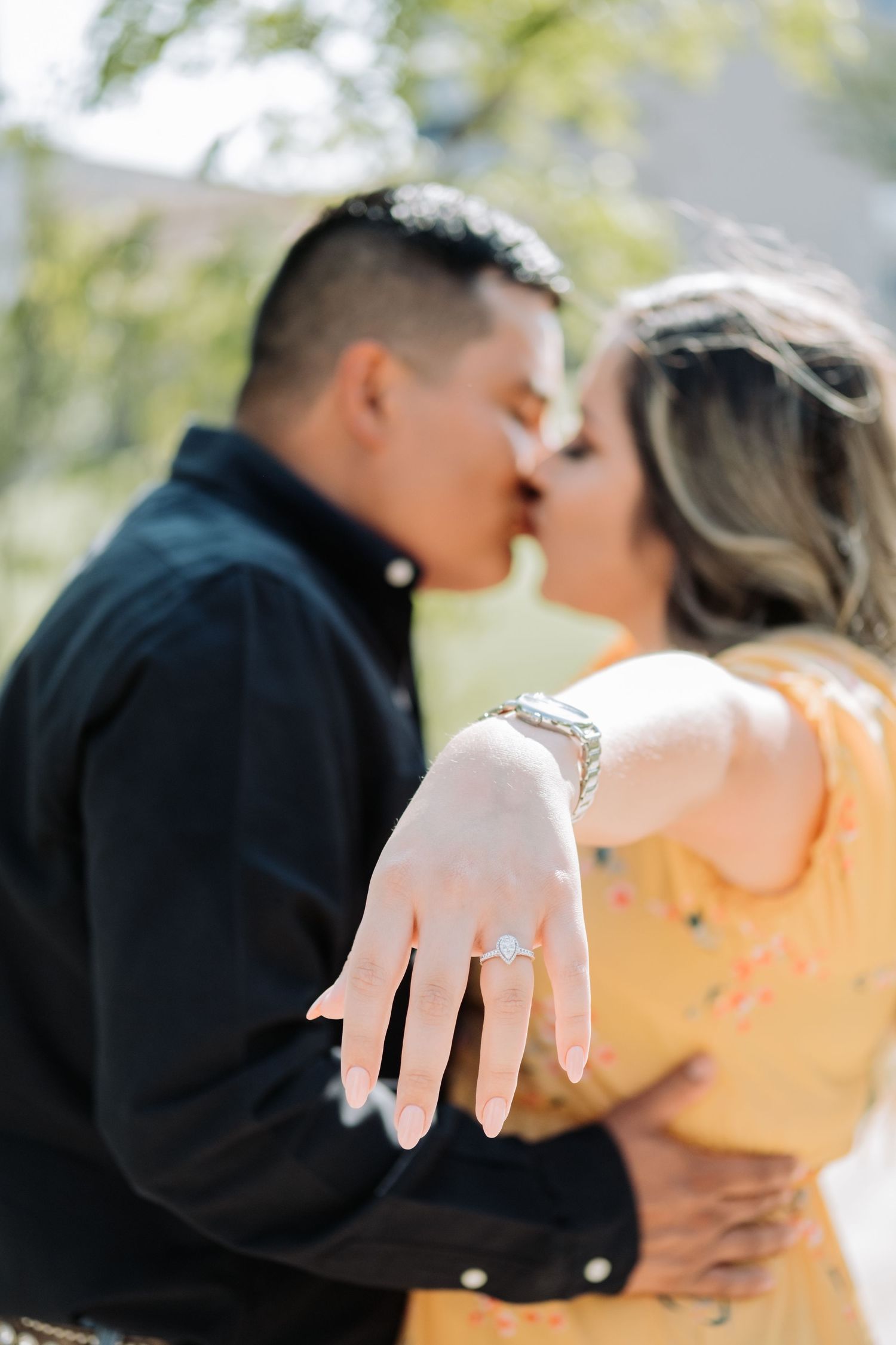 Close-up of engagement ring as couple shares kiss outdoors in sunlight.