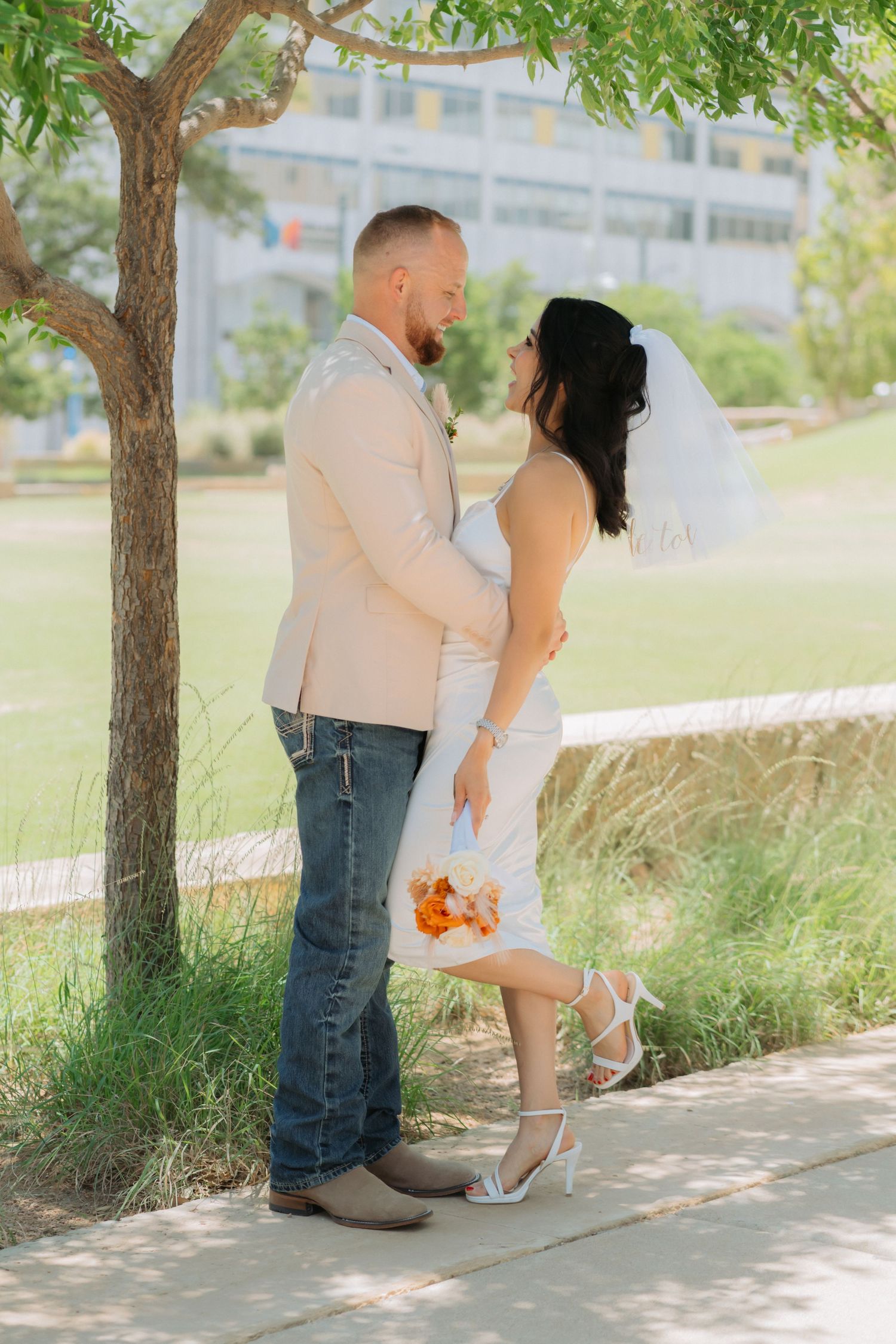 A couple in wedding attire stands beneath a tree on a paved path during a sunny outdoor photo session.