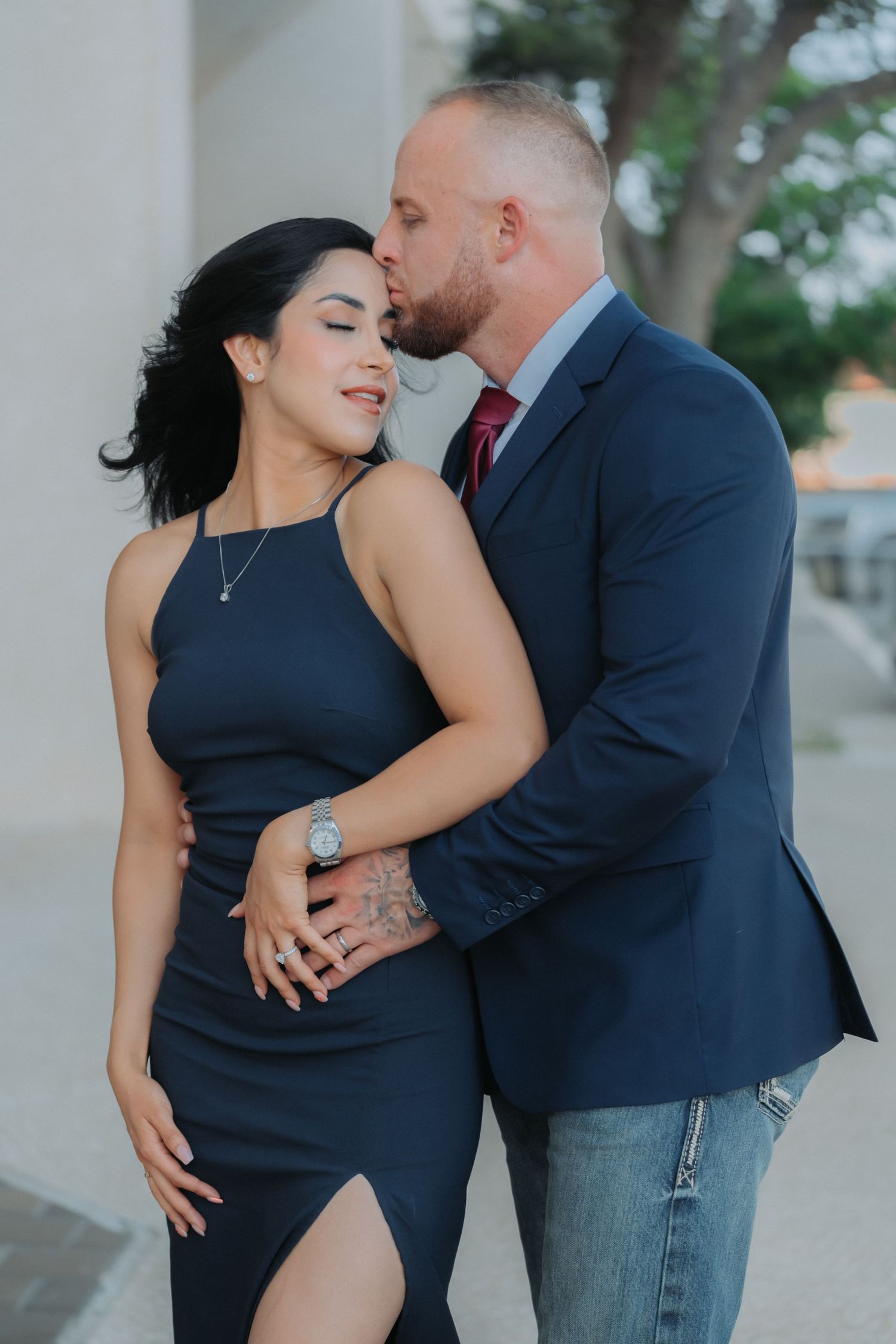 A romantic couple pose together outdoors, with her in a navy dress and him in a suit jacket and jeans.