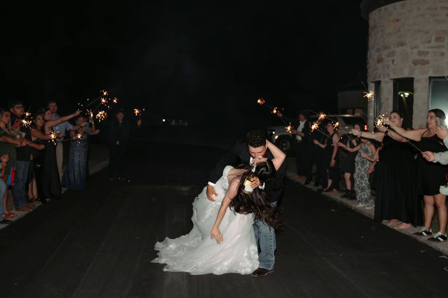 Wedding guests create a sparkler tunnel exit for the newlyweds at an outdoor nighttime celebration.