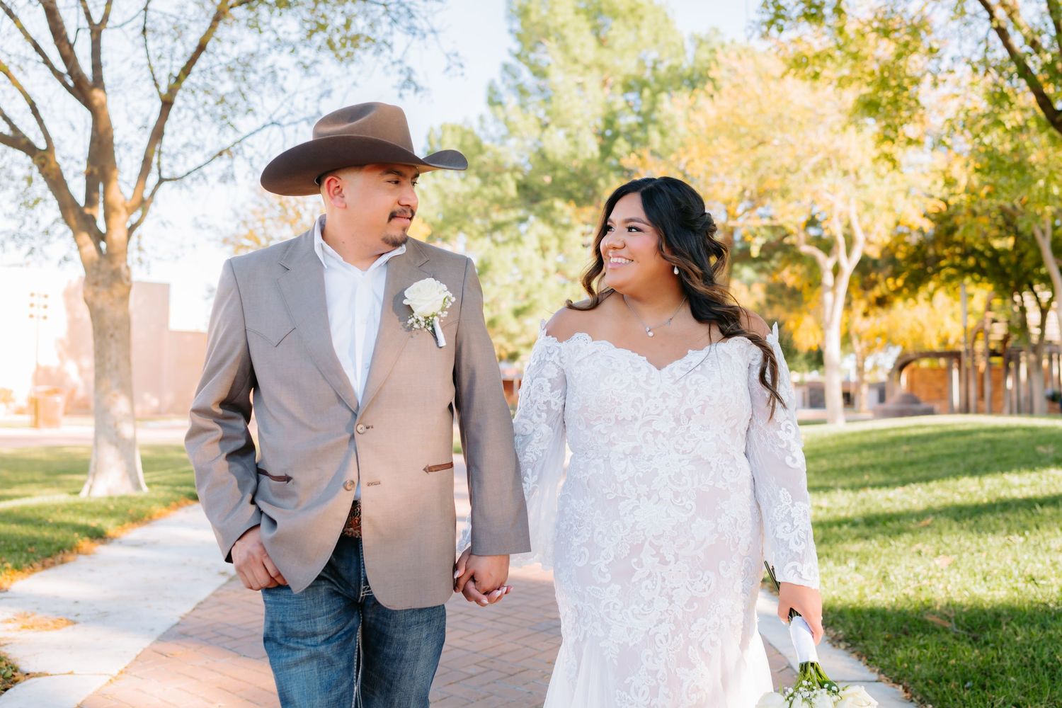 Newlyweds walk hand in hand through sunny park, bride in lace dress and groom in western attire.
