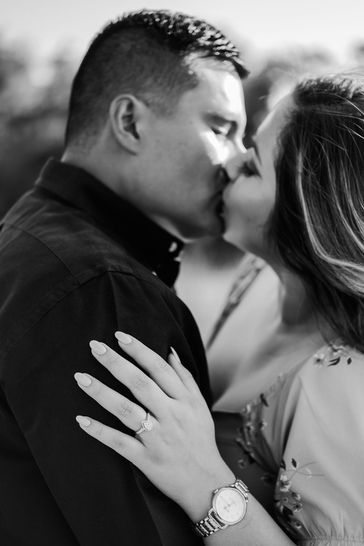 A romantic couple shares an intimate kiss as they show off an engagement ring in black and white.