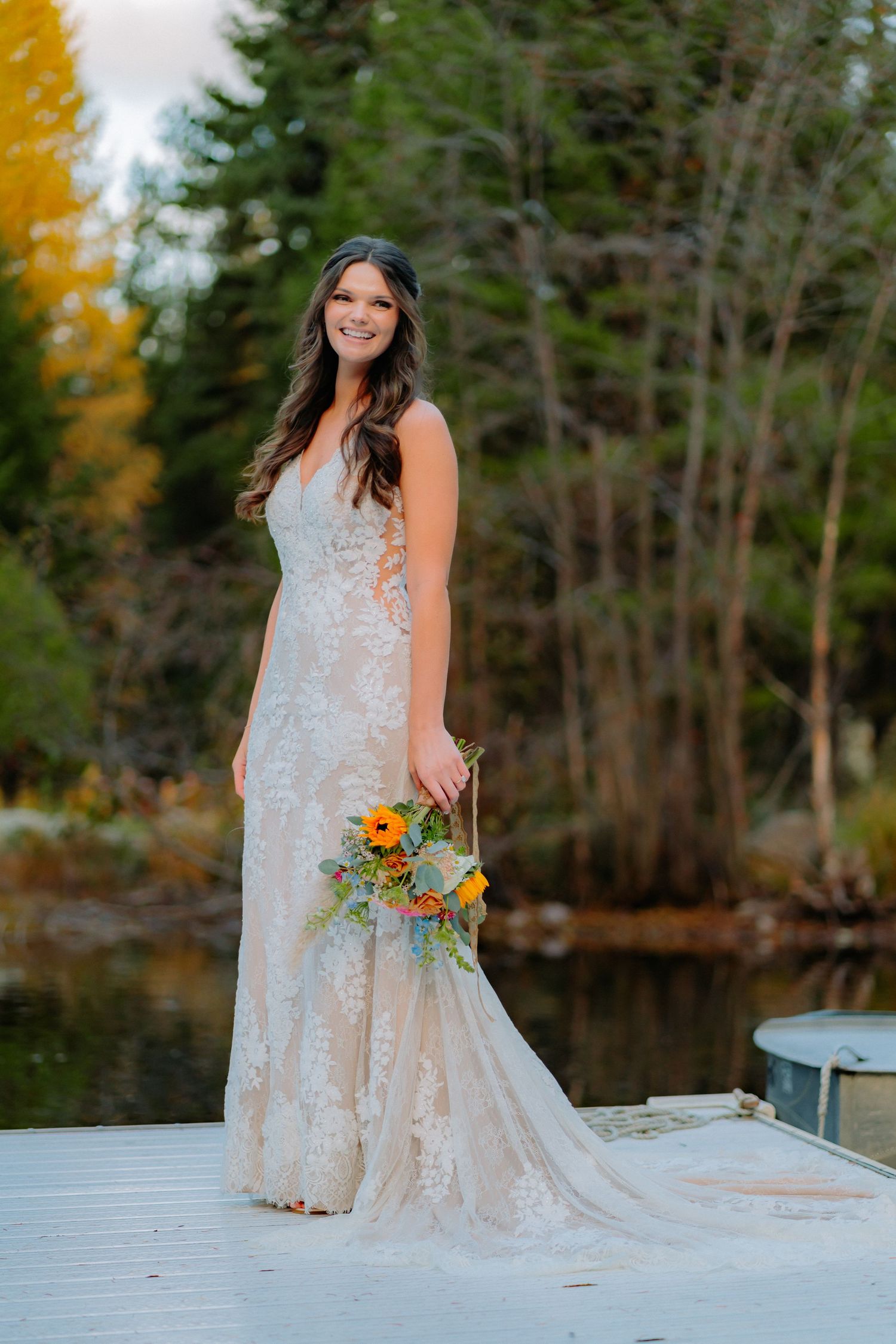 Wedding portrait of person in lace dress holding autumn bouquet on lakeside dock during fall season.