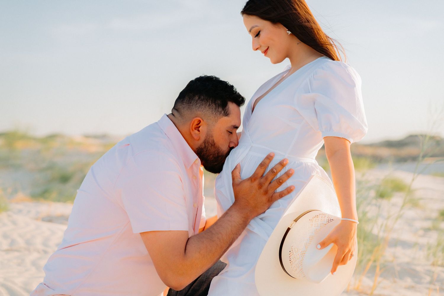 A tender maternity photo moment captured on sandy dunes during sunset.