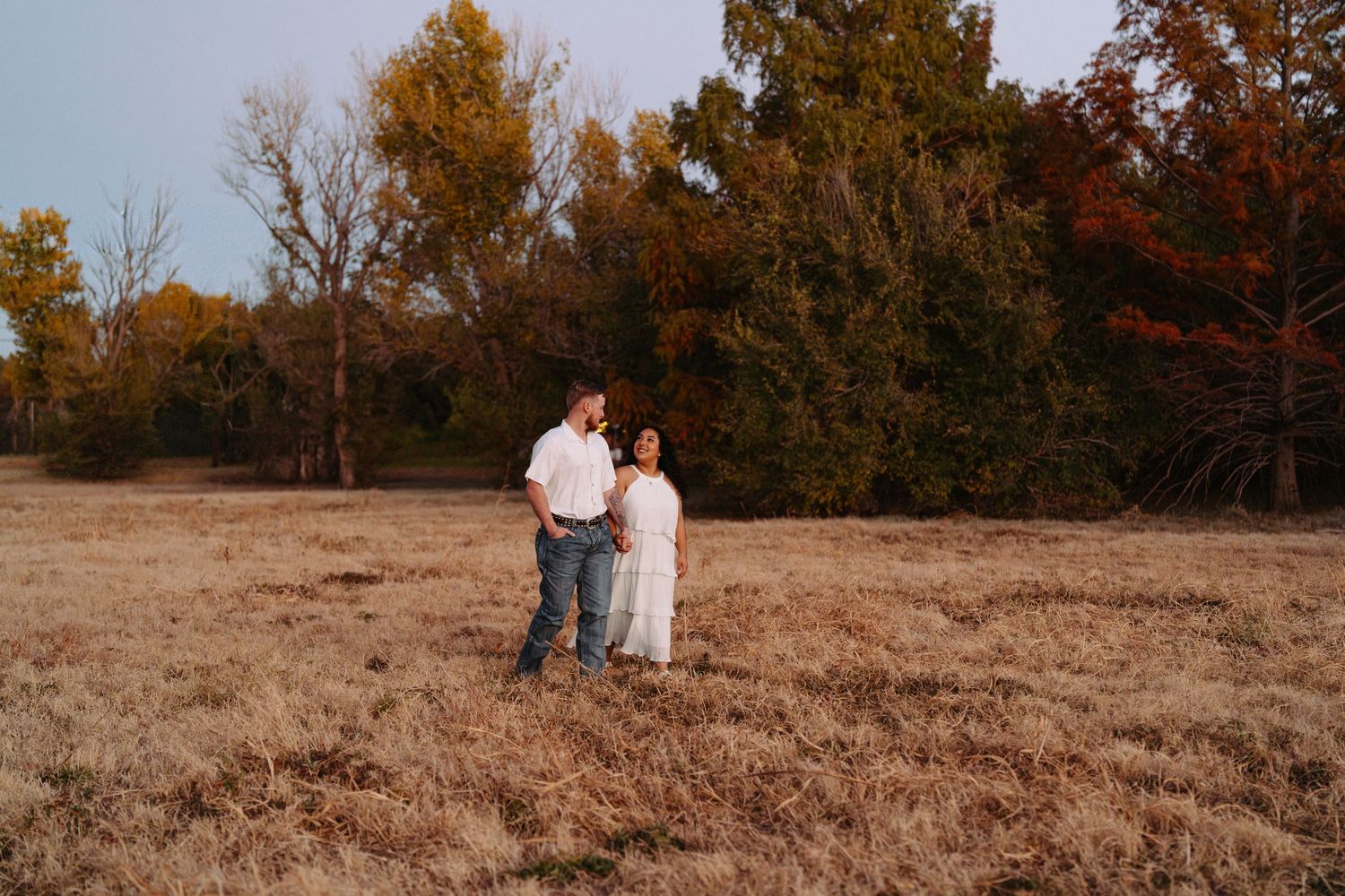 A couple walks hand in hand through a golden autumn field during sunset with fall foliage in the background.