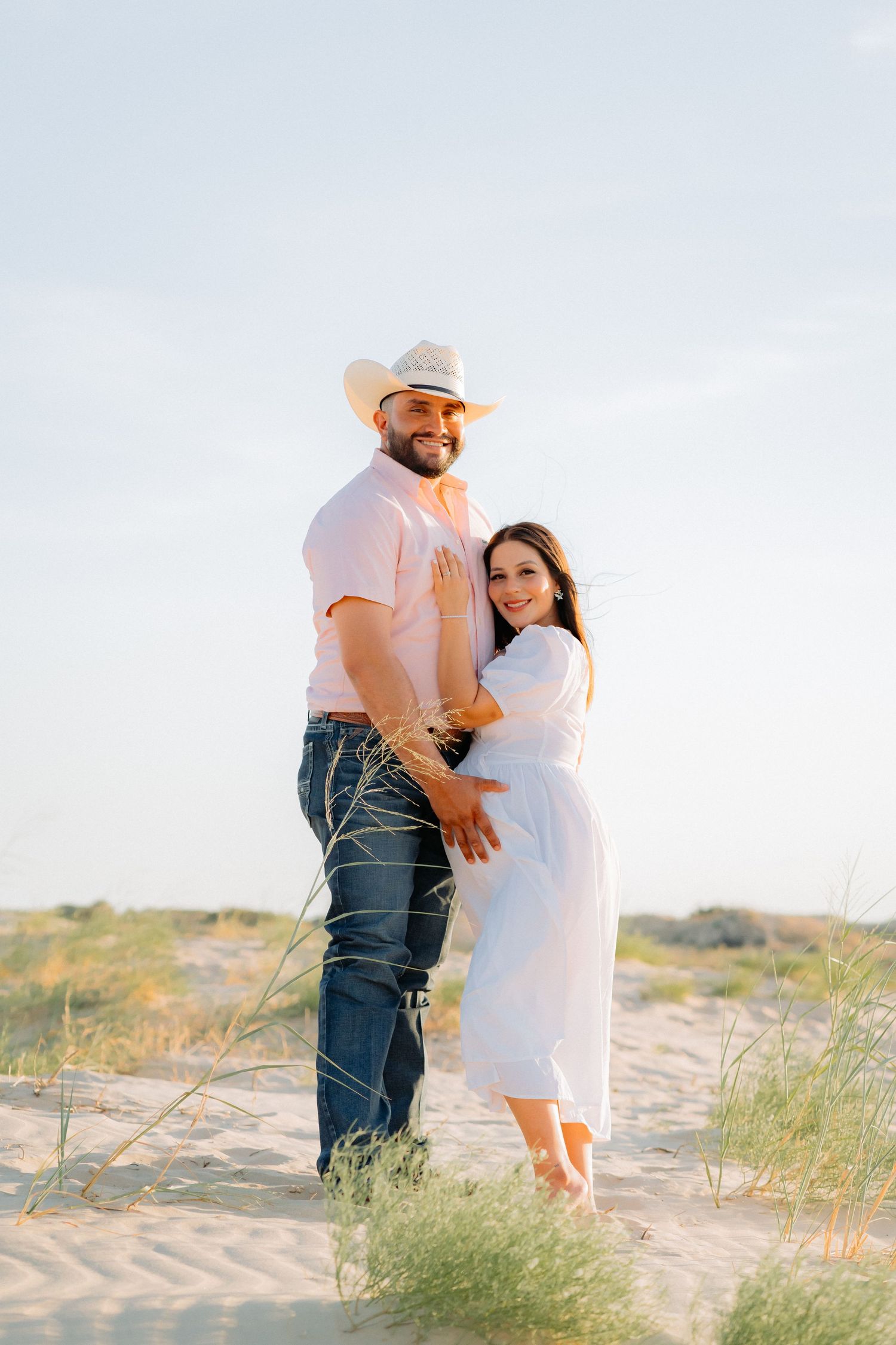 Happy couple poses for maternity photos on sandy dunes against blue sky.