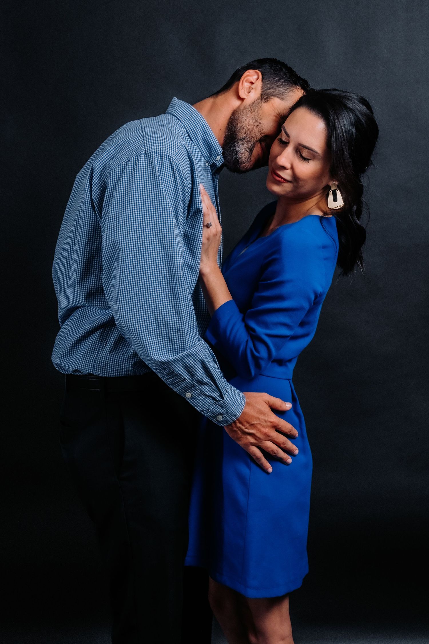 Intimate couple portrait against dark background with woman in blue dress and man in denim shirt.