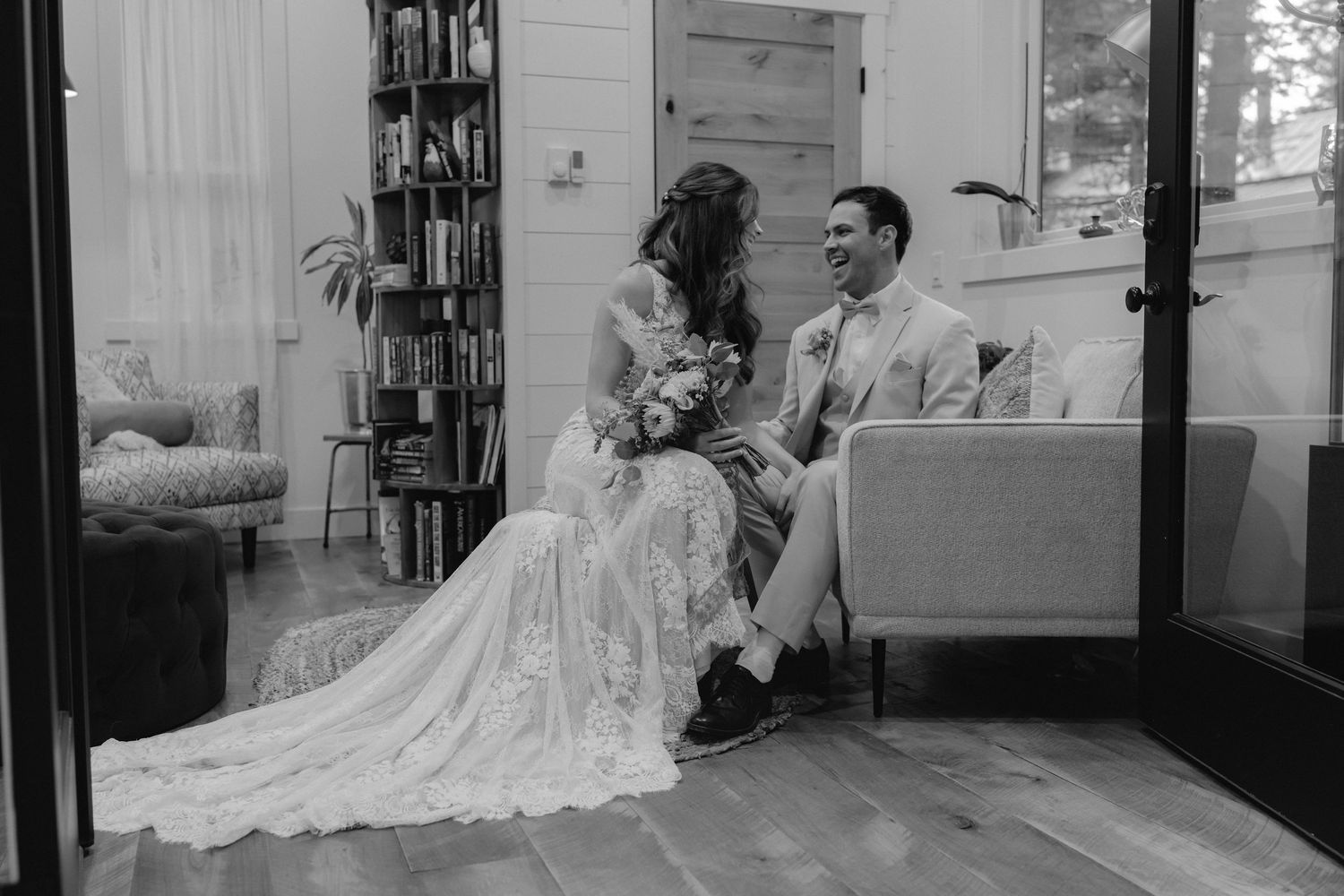 Black and white photo of couple sitting intimately on couch in wedding attire with flowing lace dress.