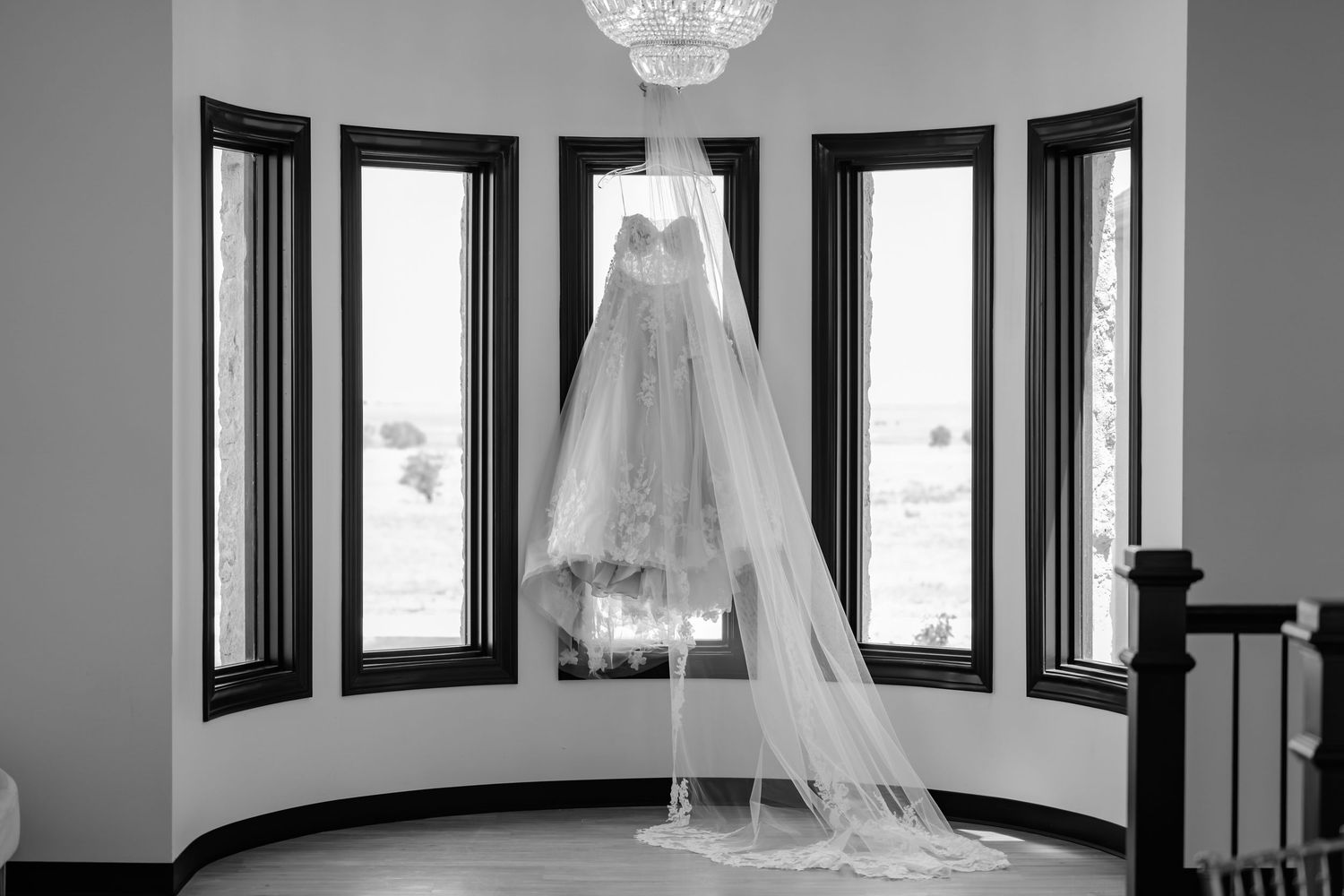 A white wedding dress hangs elegantly in a room with large windows and an ornate crystal chandelier.