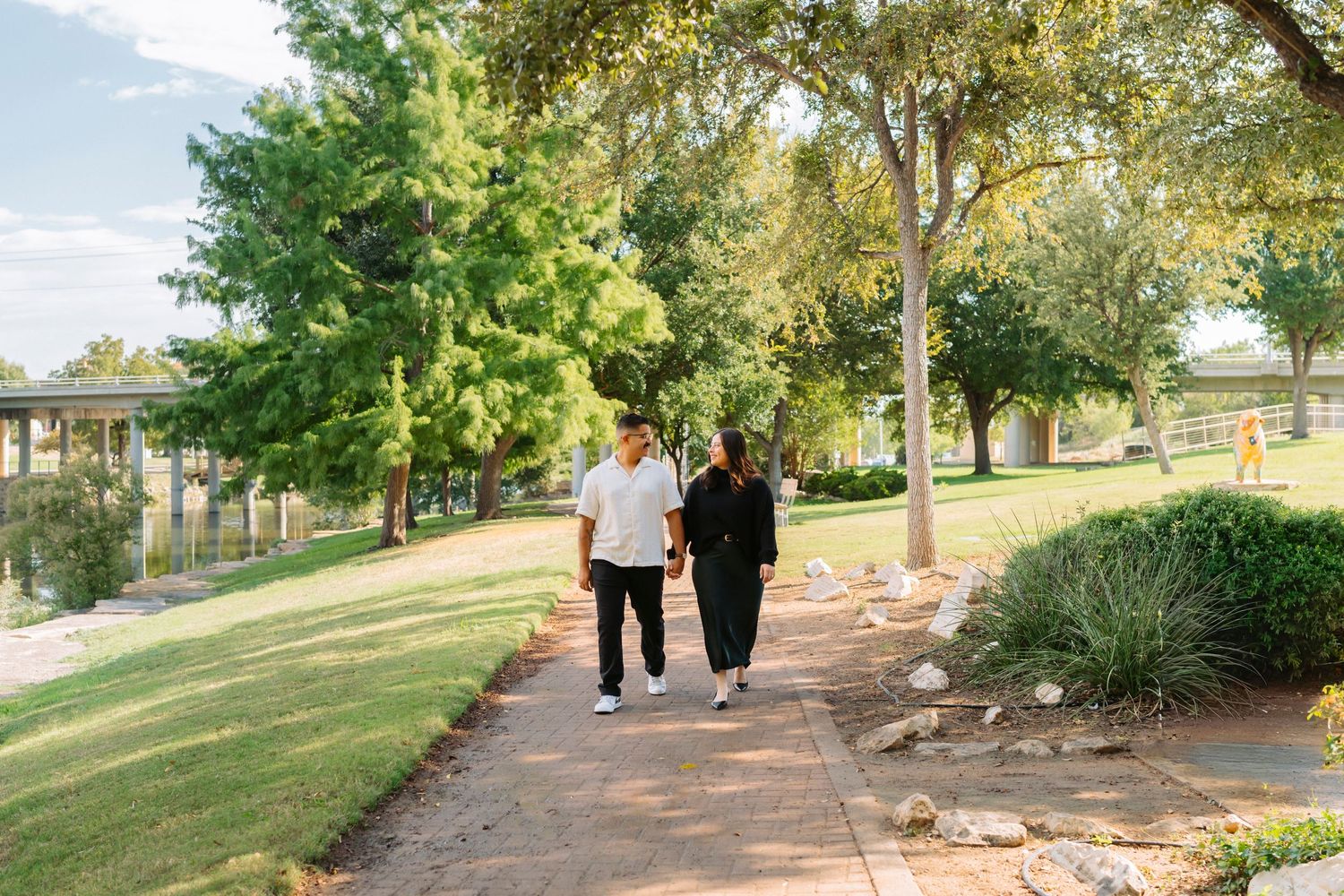 People walking along a tree-lined path in a park on a sunny day with green grass and foliage.