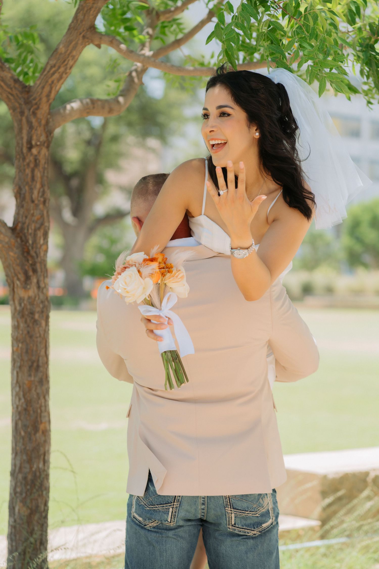 A joyful bride wearing a veil holds a bouquet of flowers while laughing in a sun-dappled outdoor garden setting.