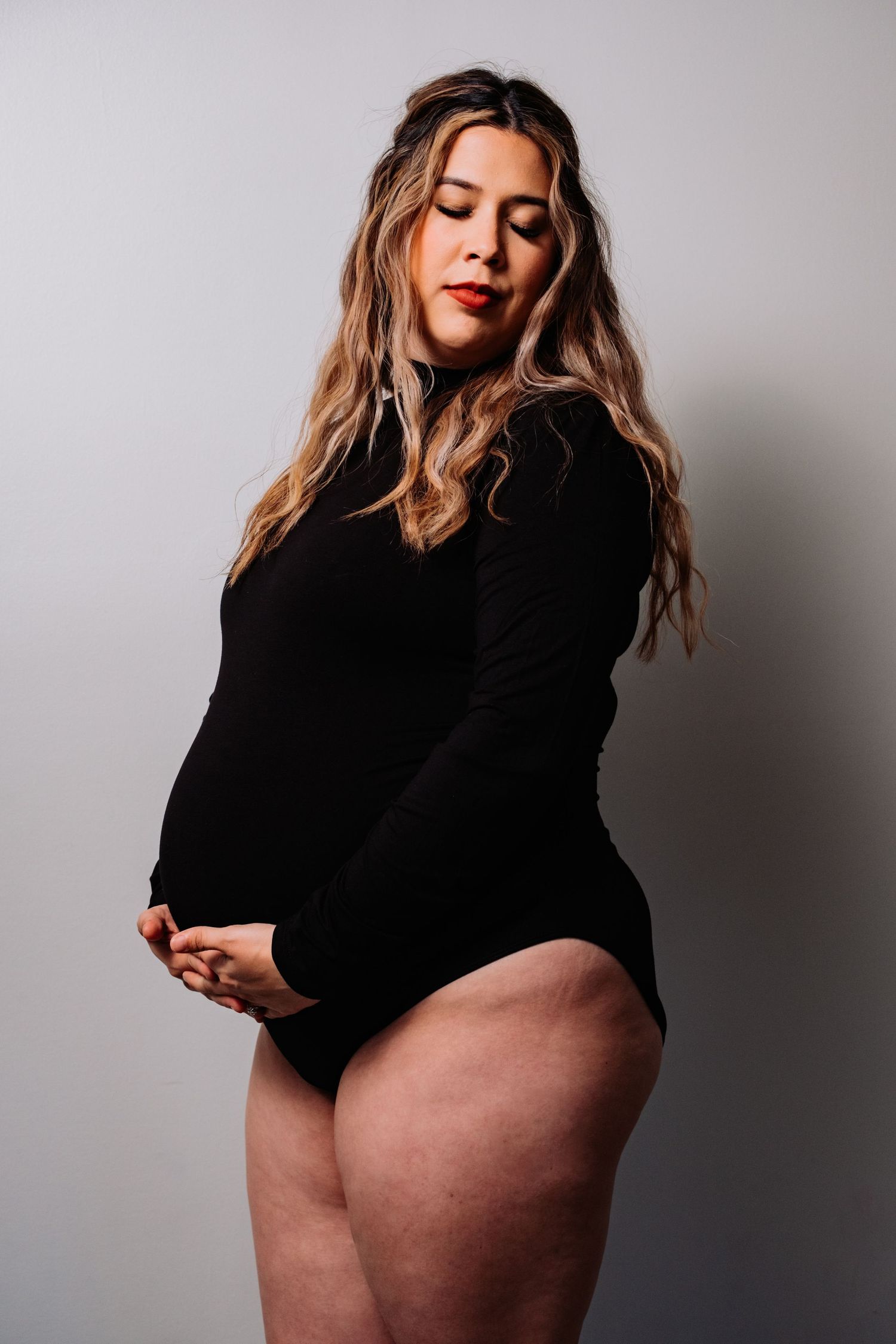 Artistic maternity portrait of woman in black bodysuit against neutral backdrop.