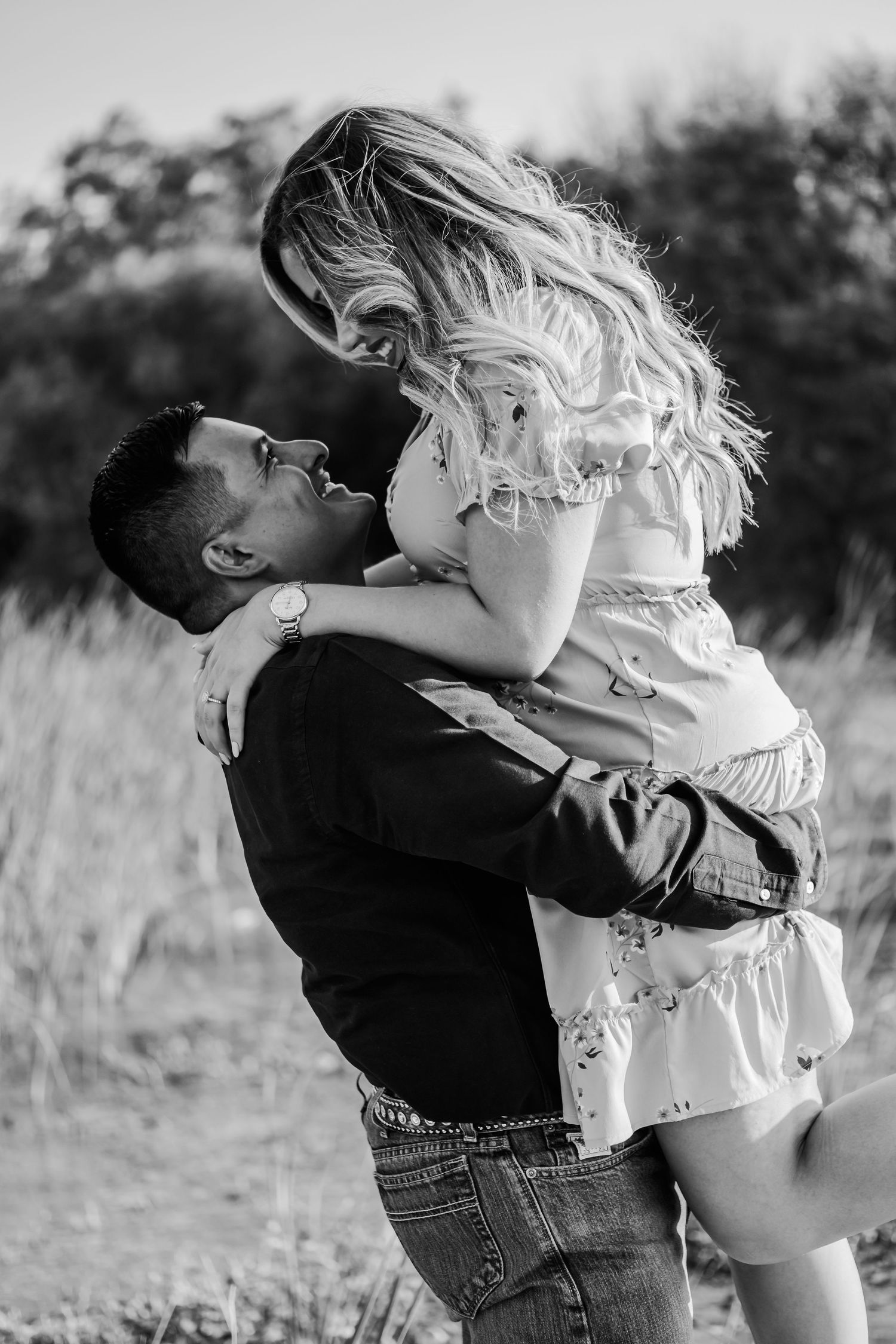 Couple shares intimate moment as man lifts woman in white dress during outdoor photoshoot.