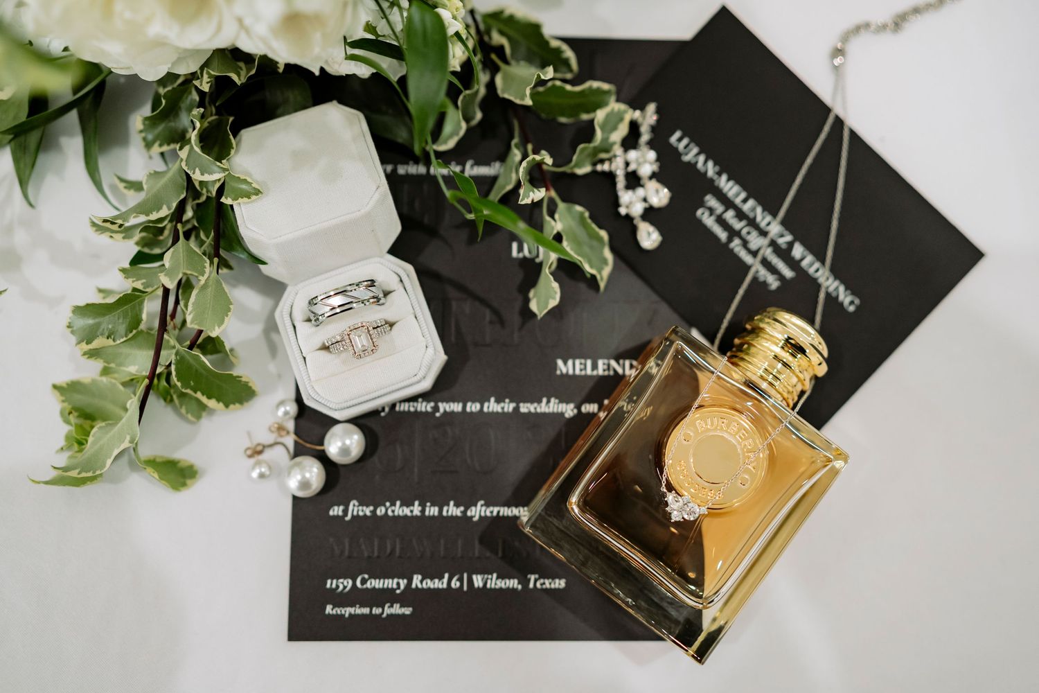 Wedding invitation and perfume bottle arranged with delicate white flowers and greenery on a light surface.