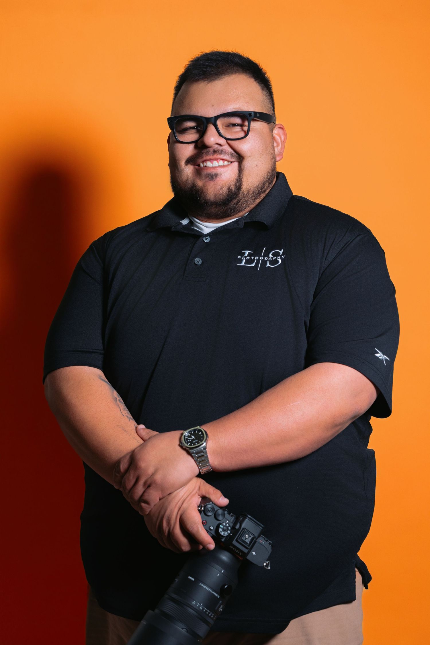 Professional photographer in black polo shirt poses with camera against vibrant orange background.