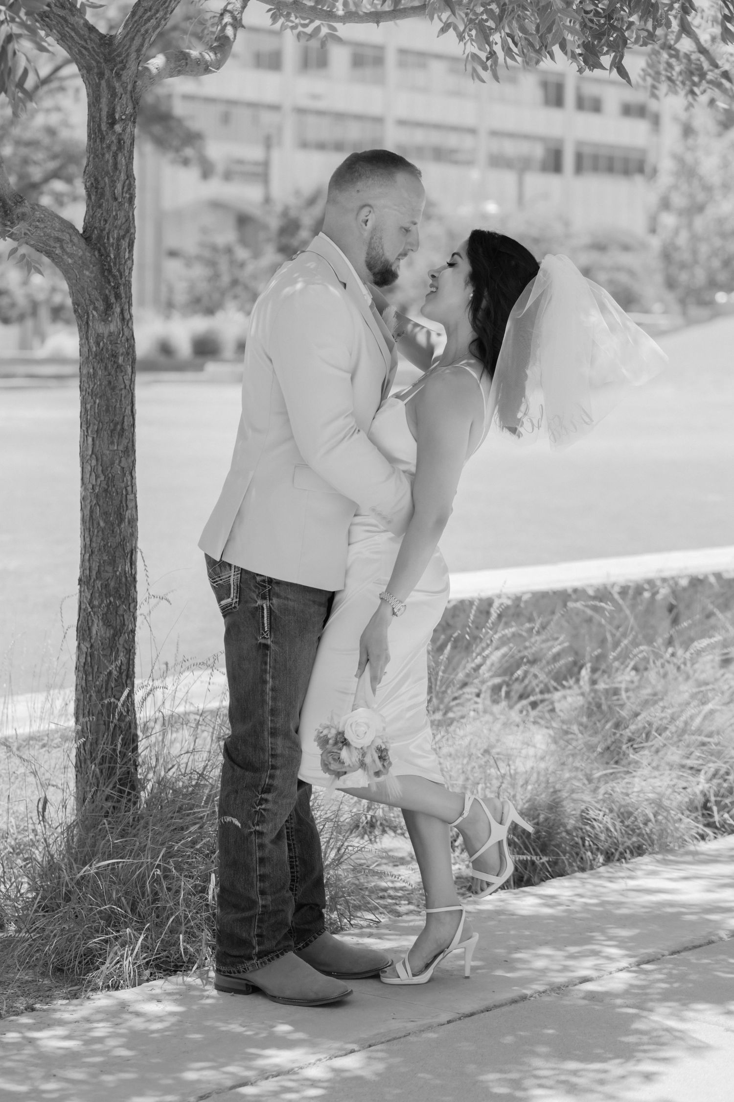 A romantic black and white wedding photo outdoors shows a loving couple embracing under a tree.