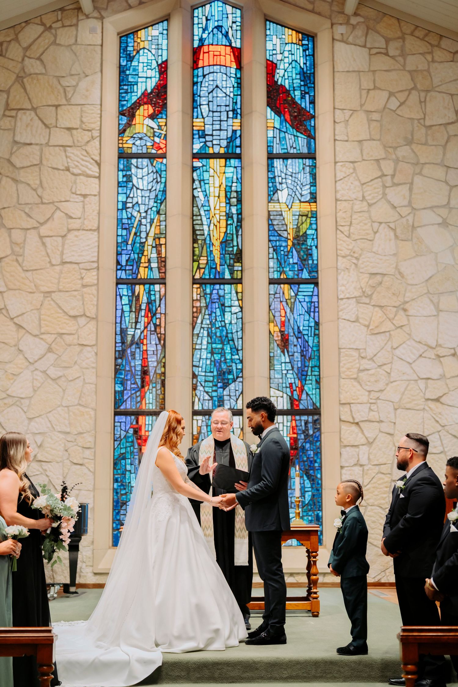 Wedding ceremony at altar with stunning blue stained glass window backdrop in stone chapel.