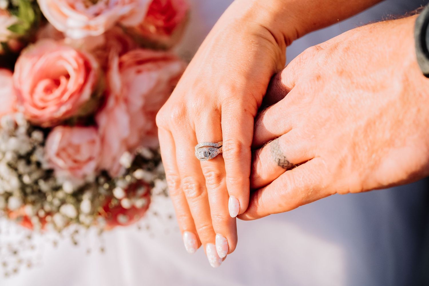 Close-up of wedding rings and hands with pink roses in background.