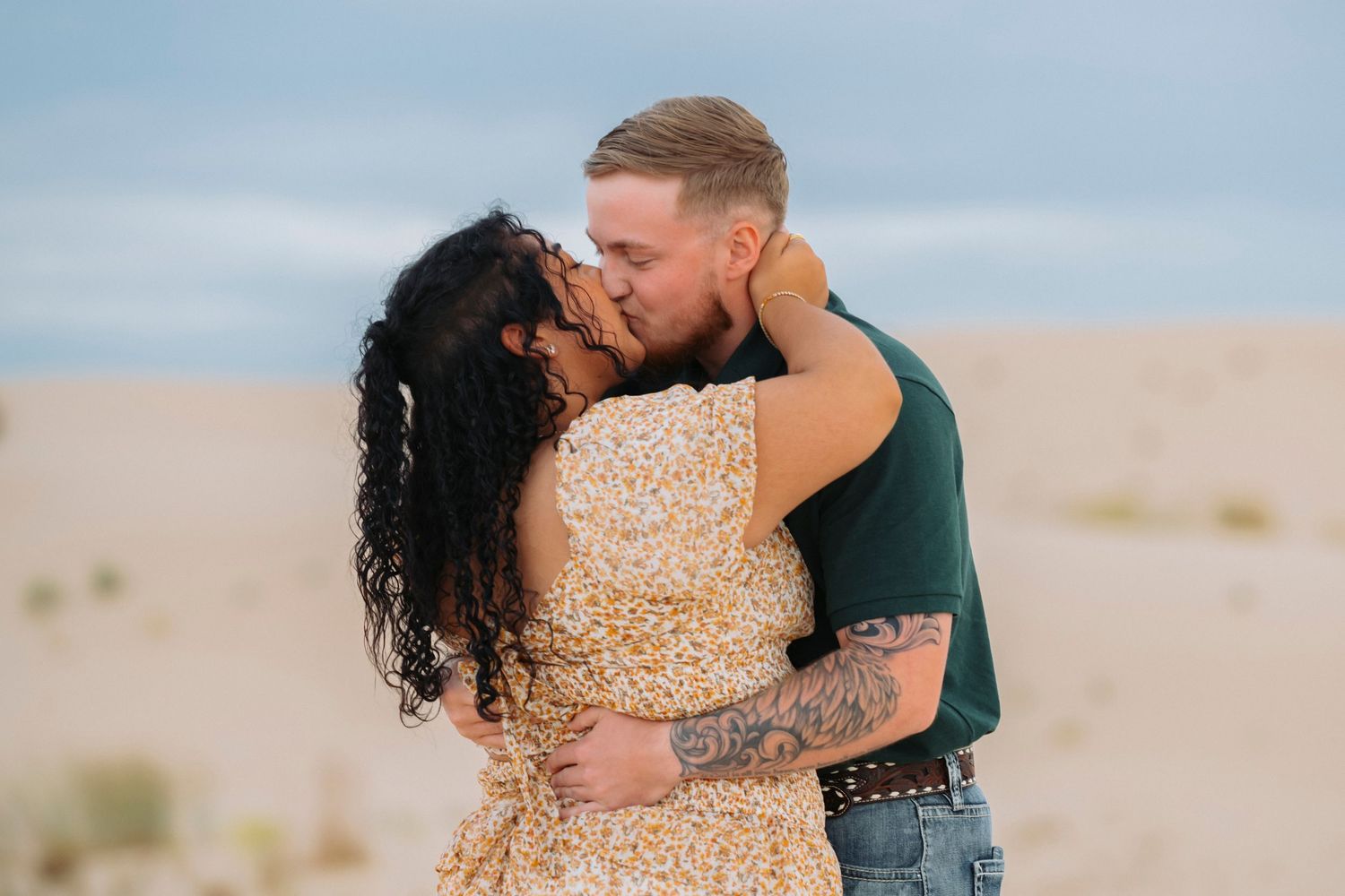 A romantic couple embraces and kisses in a desert setting during golden hour, with soft clouds in the background.